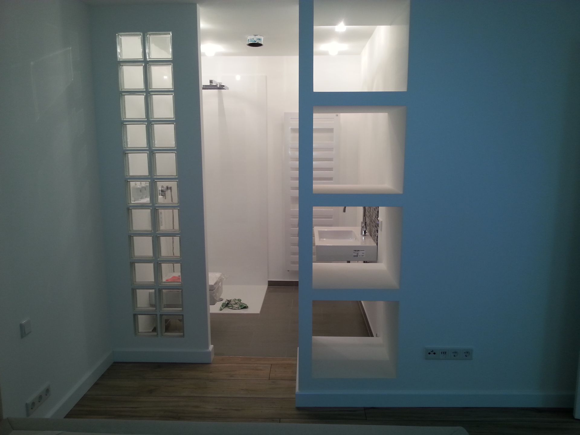 Badezimmer mit weißen Wänden, einer Glasbausteinwand und offenen Regalen. Dusche und Waschbecken sind sichtbar.