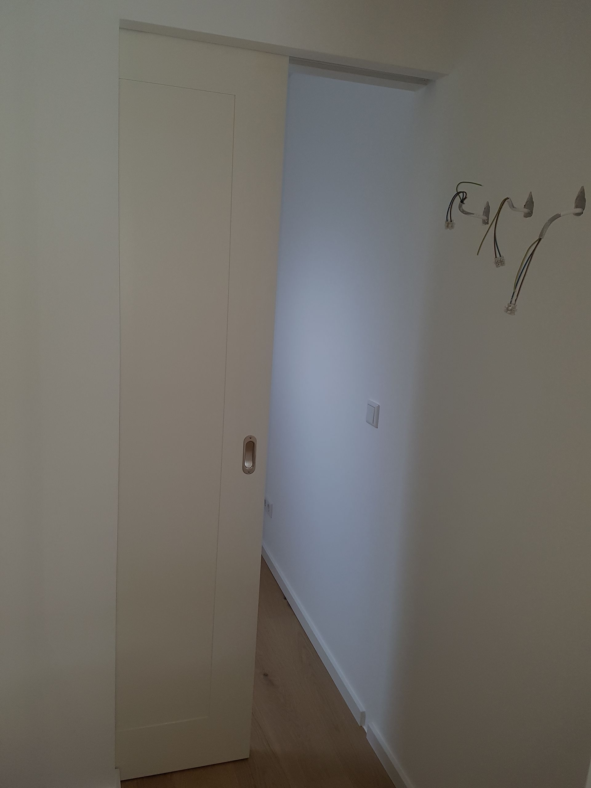 Eine weiße Schiebetür öffnet sich und gibt den Blick frei auf einen Flur mit hellem Holzboden und schlichten weißen Wänden.
