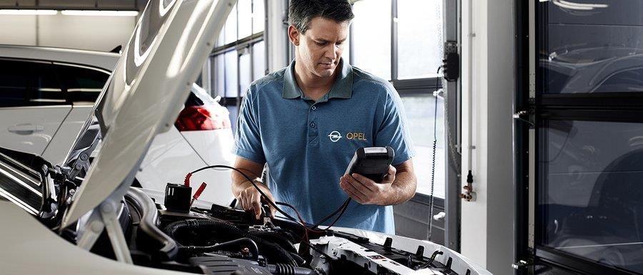 Ein Mann arbeitet in einer Werkstatt an einem Auto und hält dabei ein Mobiltelefon in der Hand.