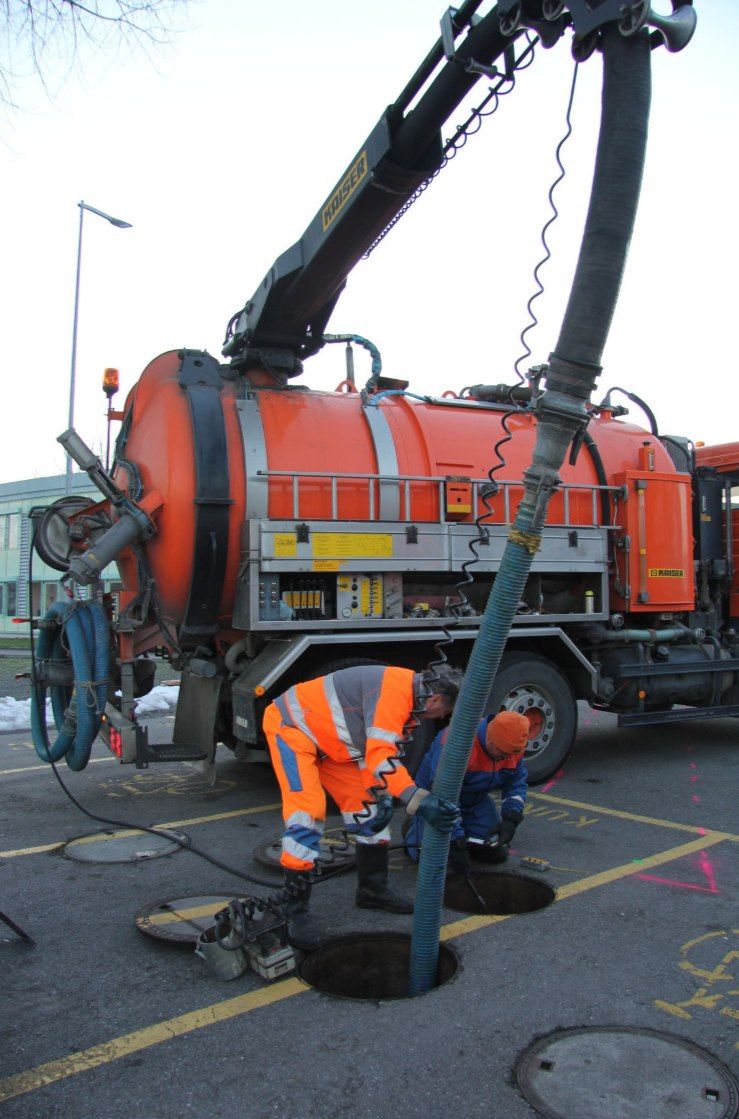Arbeiter reinigen mit einem LKW und einem Schlauch einen Entwässerungsbereich in der Nähe eines Gewässers.