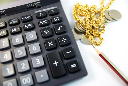Calculadora, collar de oro, monedas y lápiz sobre una superficie blanca, sugiriendo planificación o cálculos financieros.