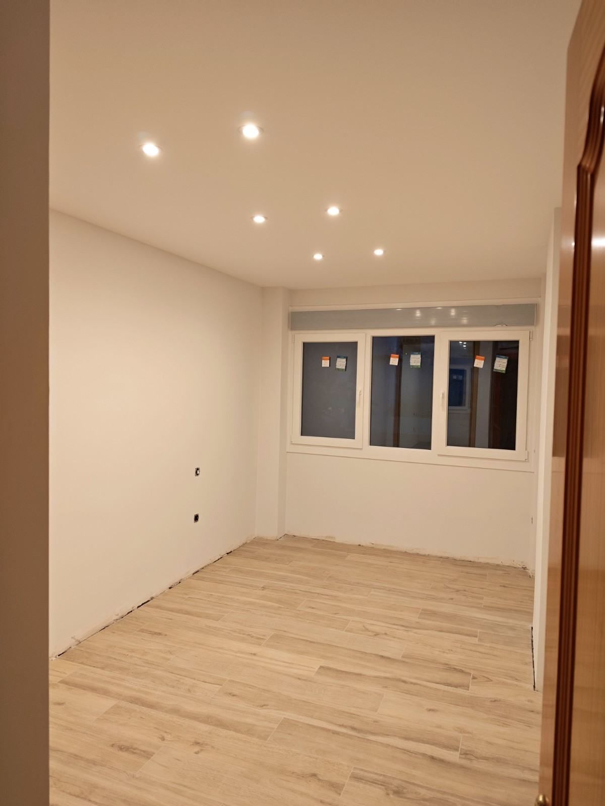 Habitación blanca vacía con suelo de madera clara, luces empotradas en el techo y una ventana al fondo.