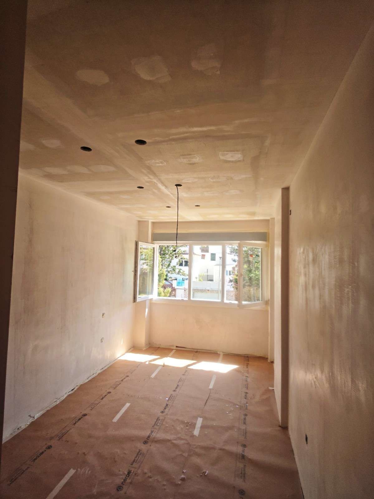 Habitación vacía sin terminar, con paredes blancas, parches de techo a la vista y luz solar que entra por una ventana.