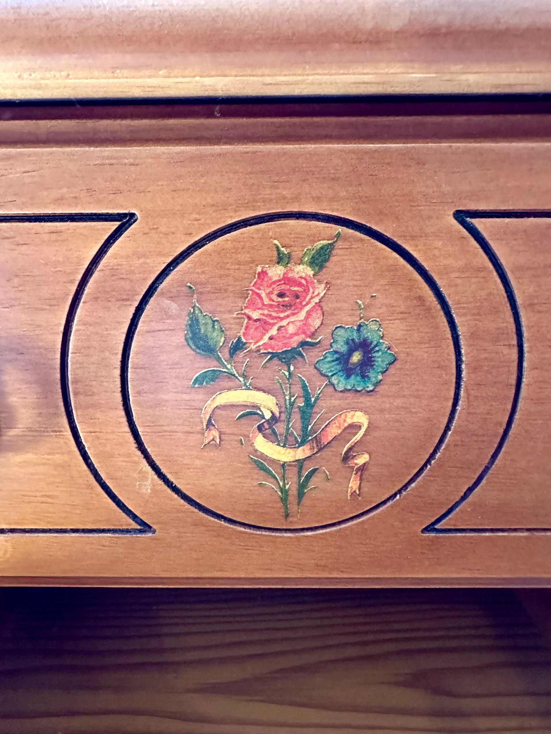 Detalle de tocador de madera con incrustaciones florales: una rosa rosa, una flor azul, hojas verdes y una cinta dorada en un círculo.
