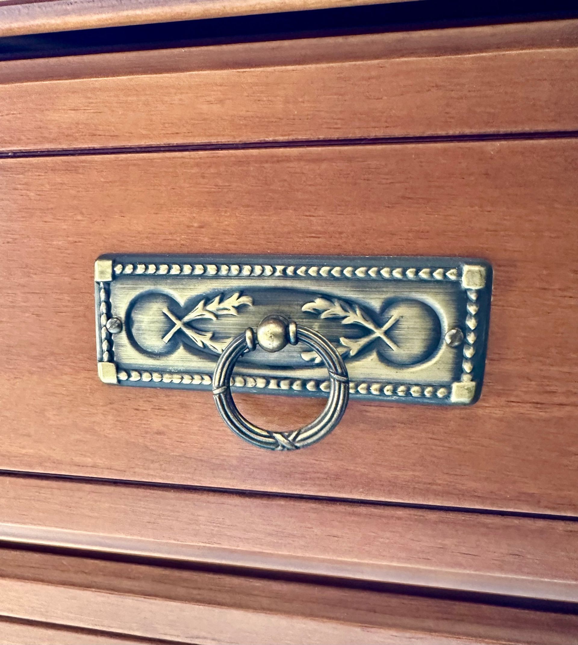 Tirador de latón para cajón de una cómoda de madera. Presenta un tirador de anilla y un diseño decorativo de hojas.