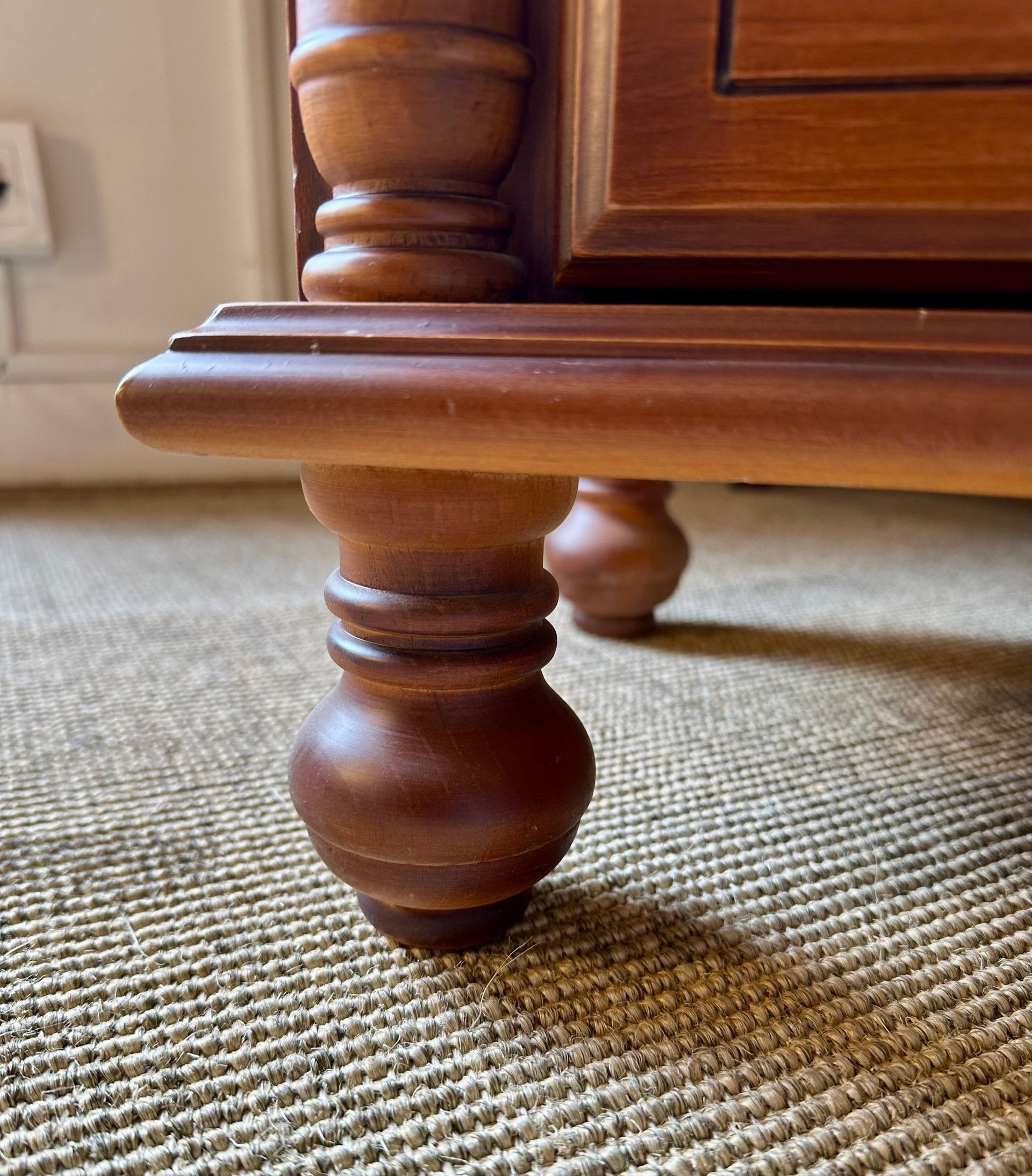 Pata de mueble de madera sobre una alfombra marrón texturizada, que muestra detalles de madera torneada y sombra.
