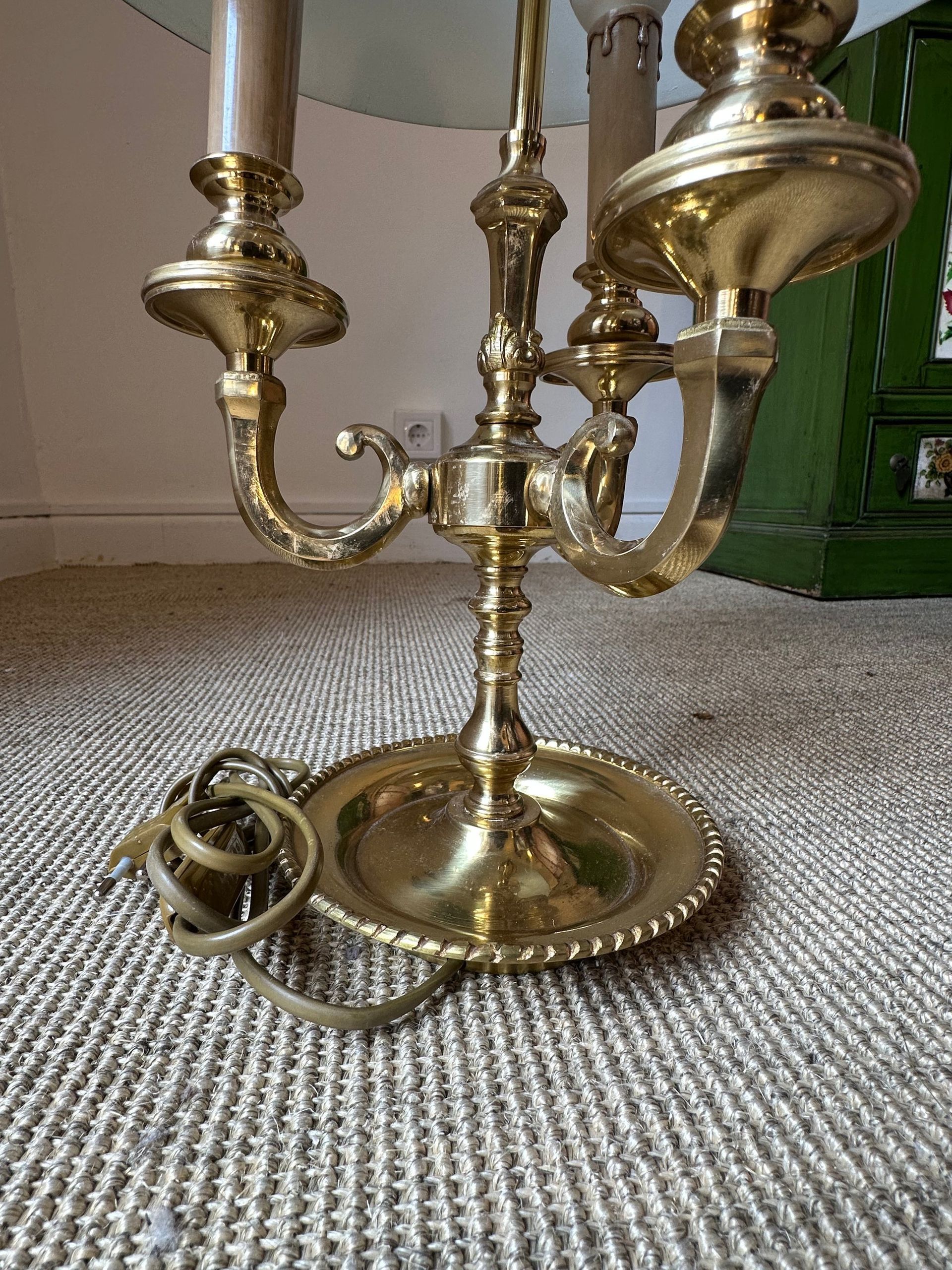 Lámpara estilo candelabro de latón sobre alfombra tejida, con un cordón en primer plano y un mueble verde al fondo.