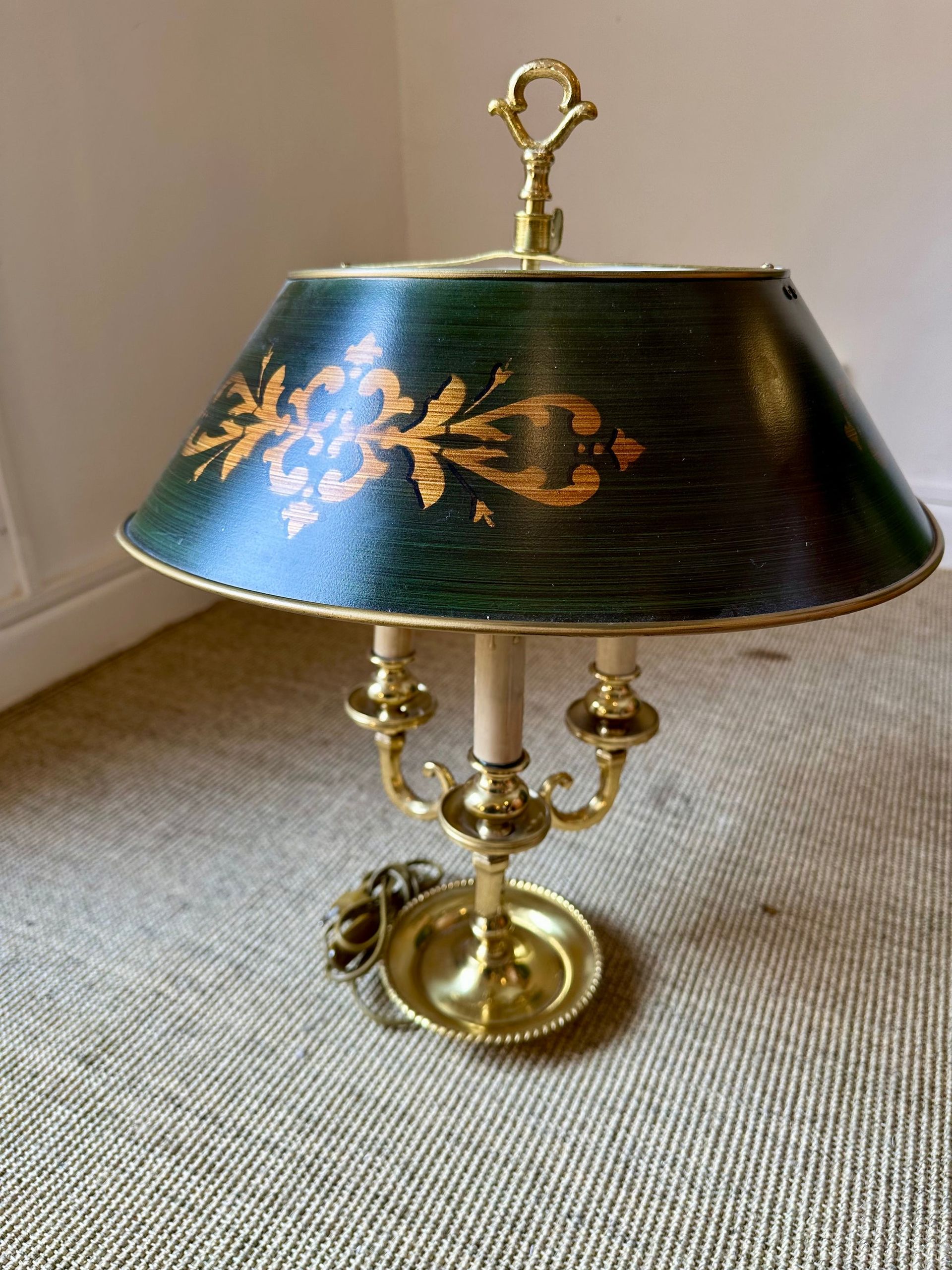 Lámpara de latón con pantalla verde que presenta detalles florales dorados sobre una alfombra neutra.