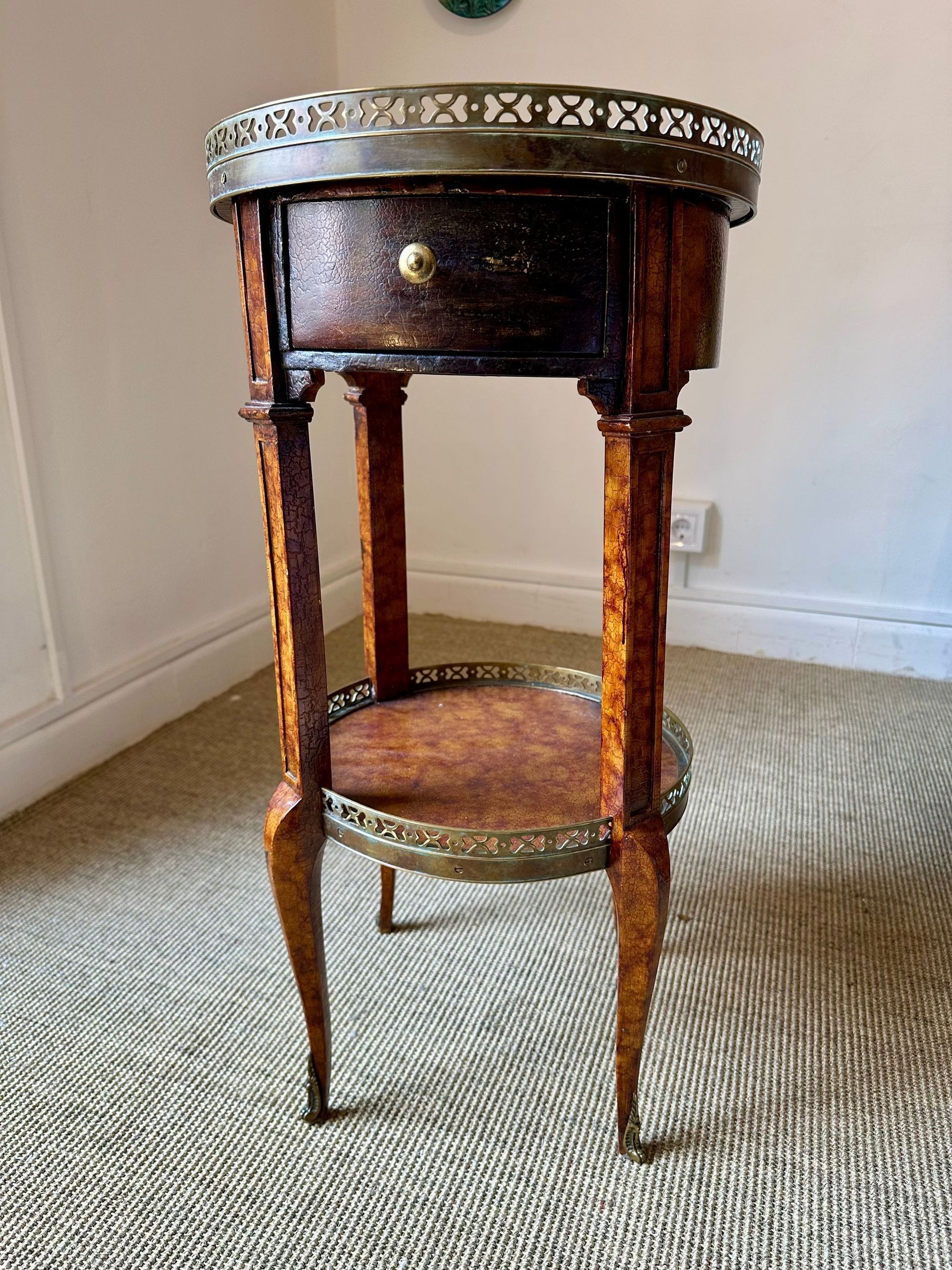 Mesa auxiliar antigua de madera con detalles de latón y un cajón, colocada sobre una alfombra estampada.