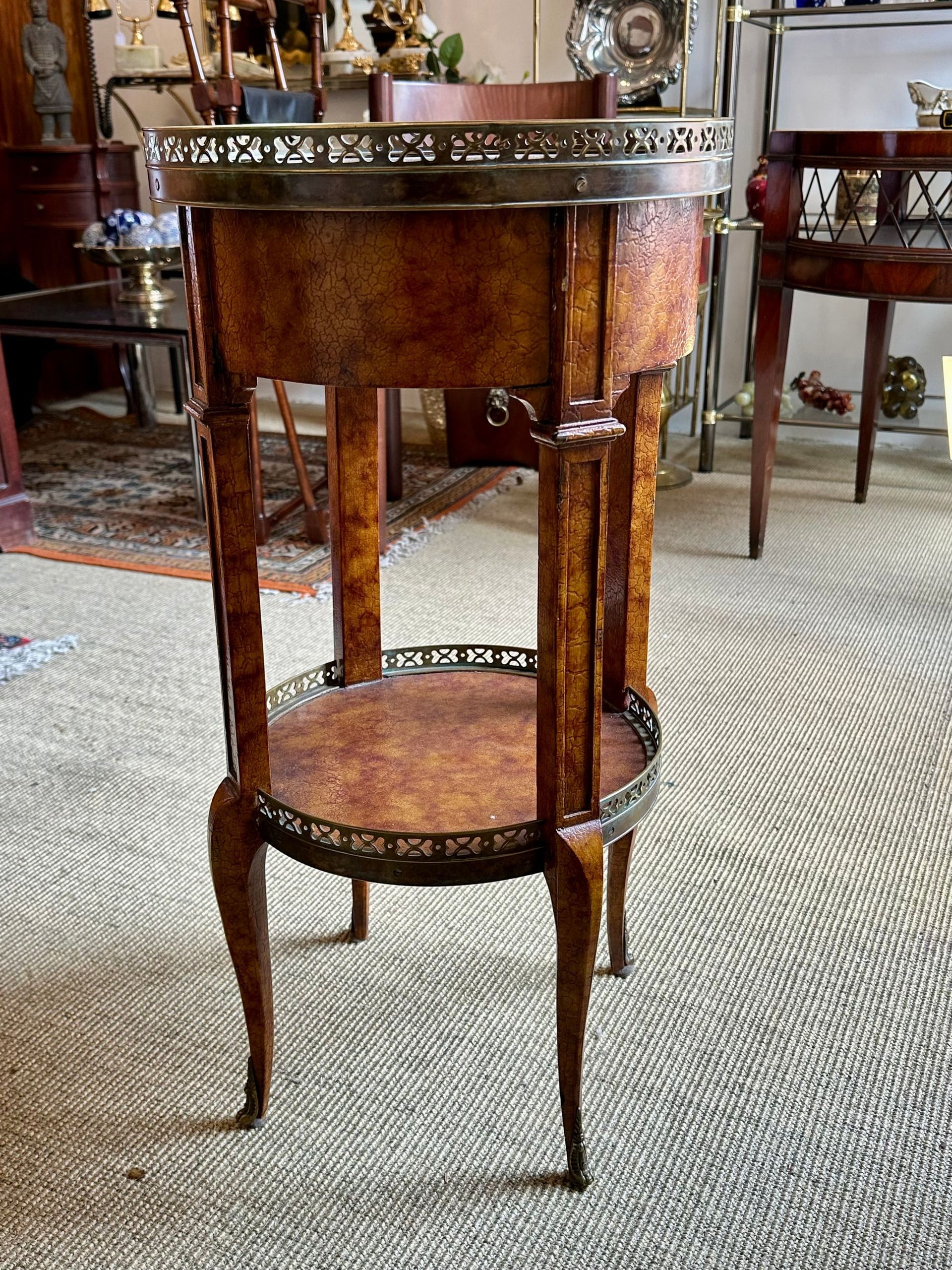 Mesa auxiliar alta de color marrón y dorado con borde decorativo y estante inferior, colocada sobre una alfombra clara.