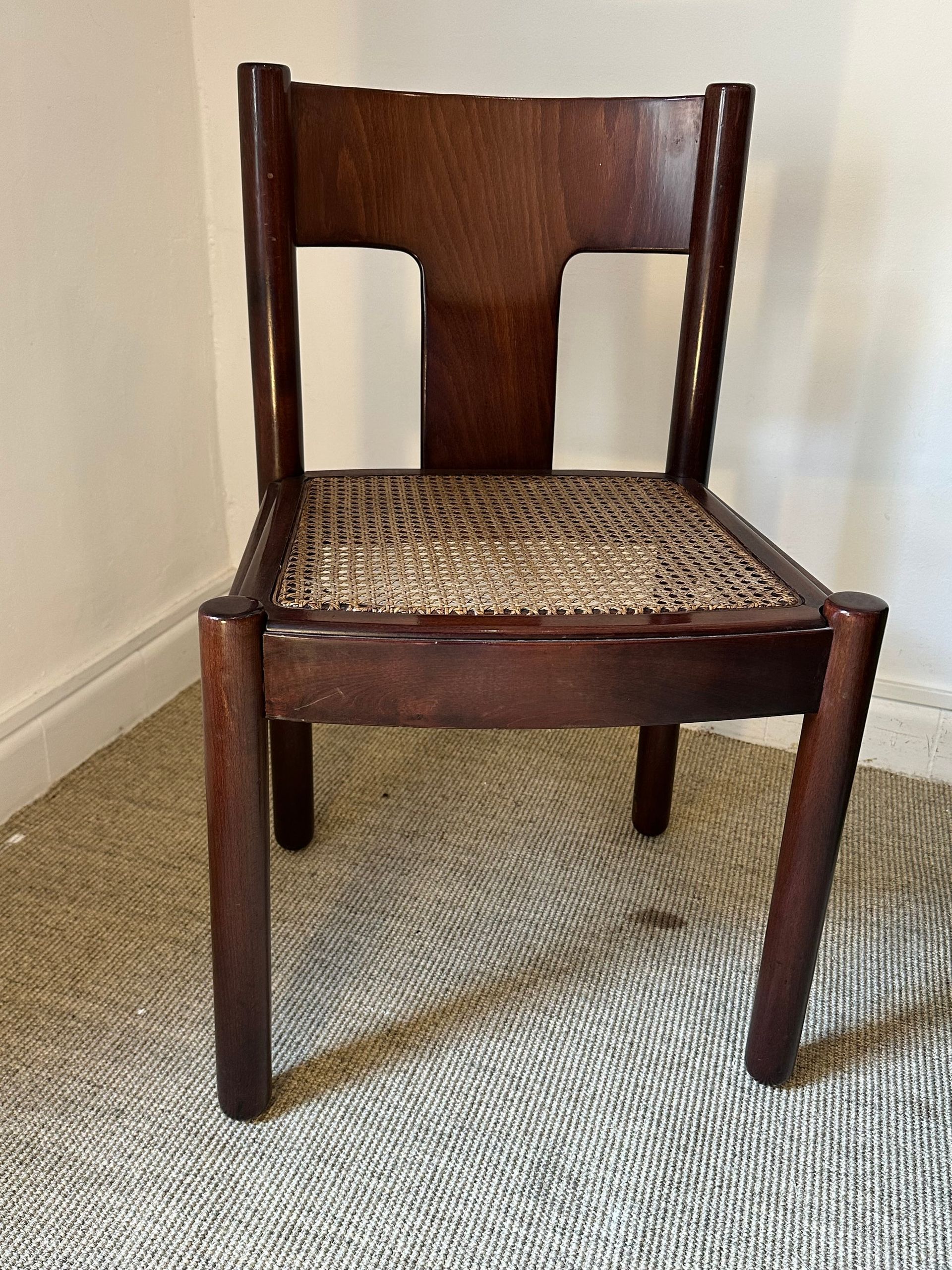 Silla de madera marrón con asiento tejido, contra una pared sobre una alfombra estampada.