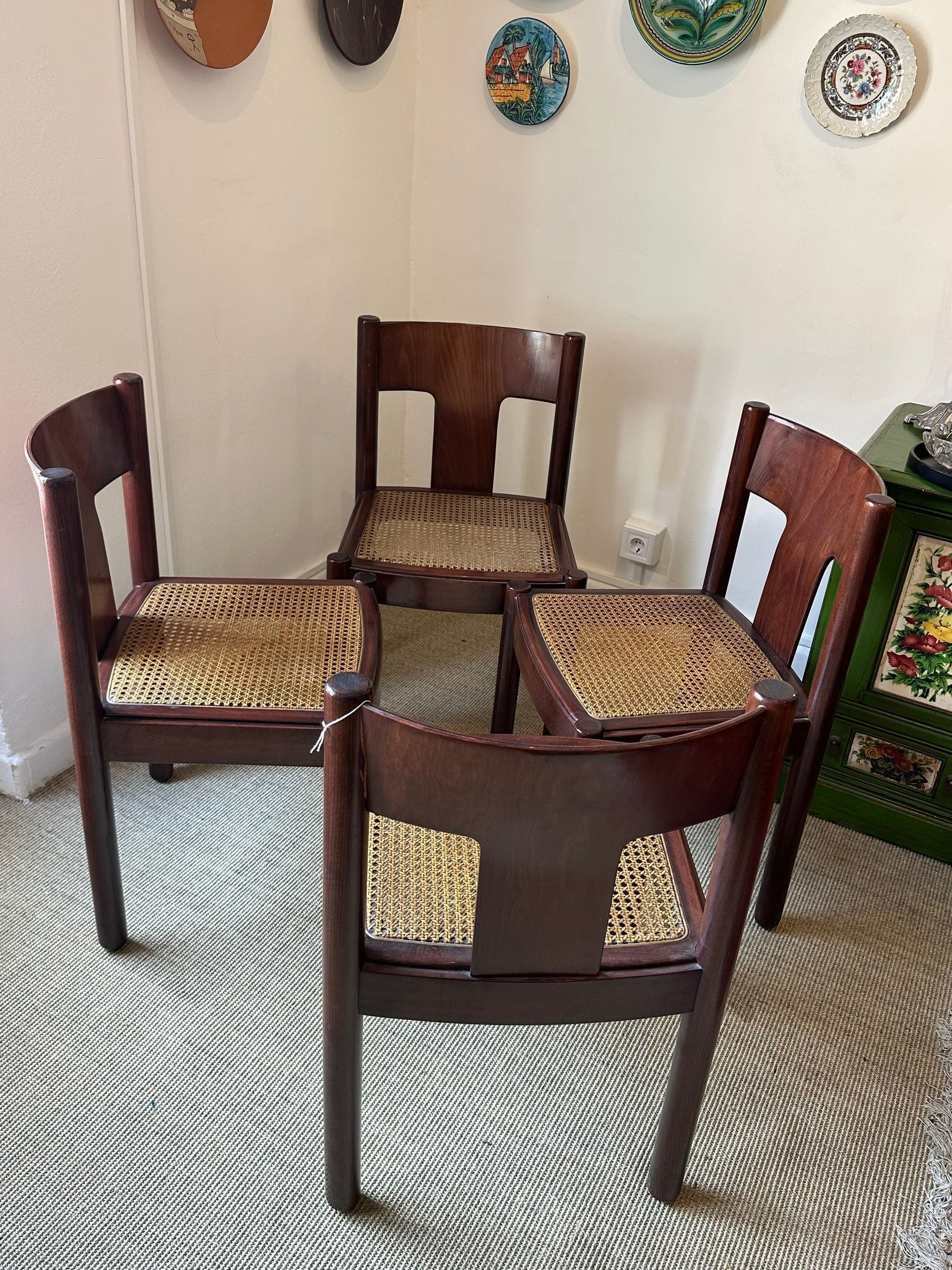 Cuatro sillas de madera oscura con asientos tejidos sobre una alfombra estampada. Platos decorativos en la pared.