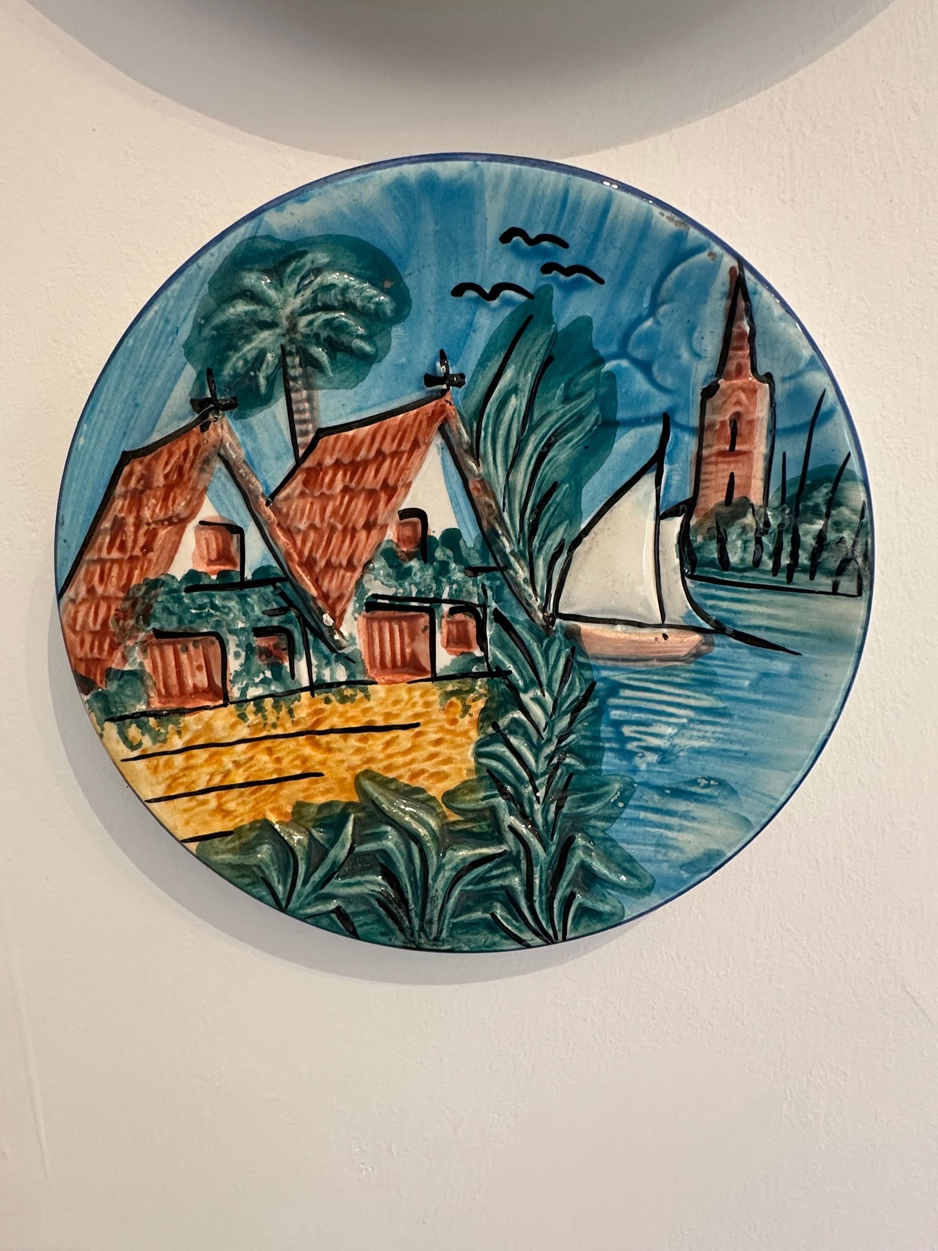 Plato decorativo con escena costera: cielo azul, palmeras, edificios con tejados rojos, un velero y una torre.