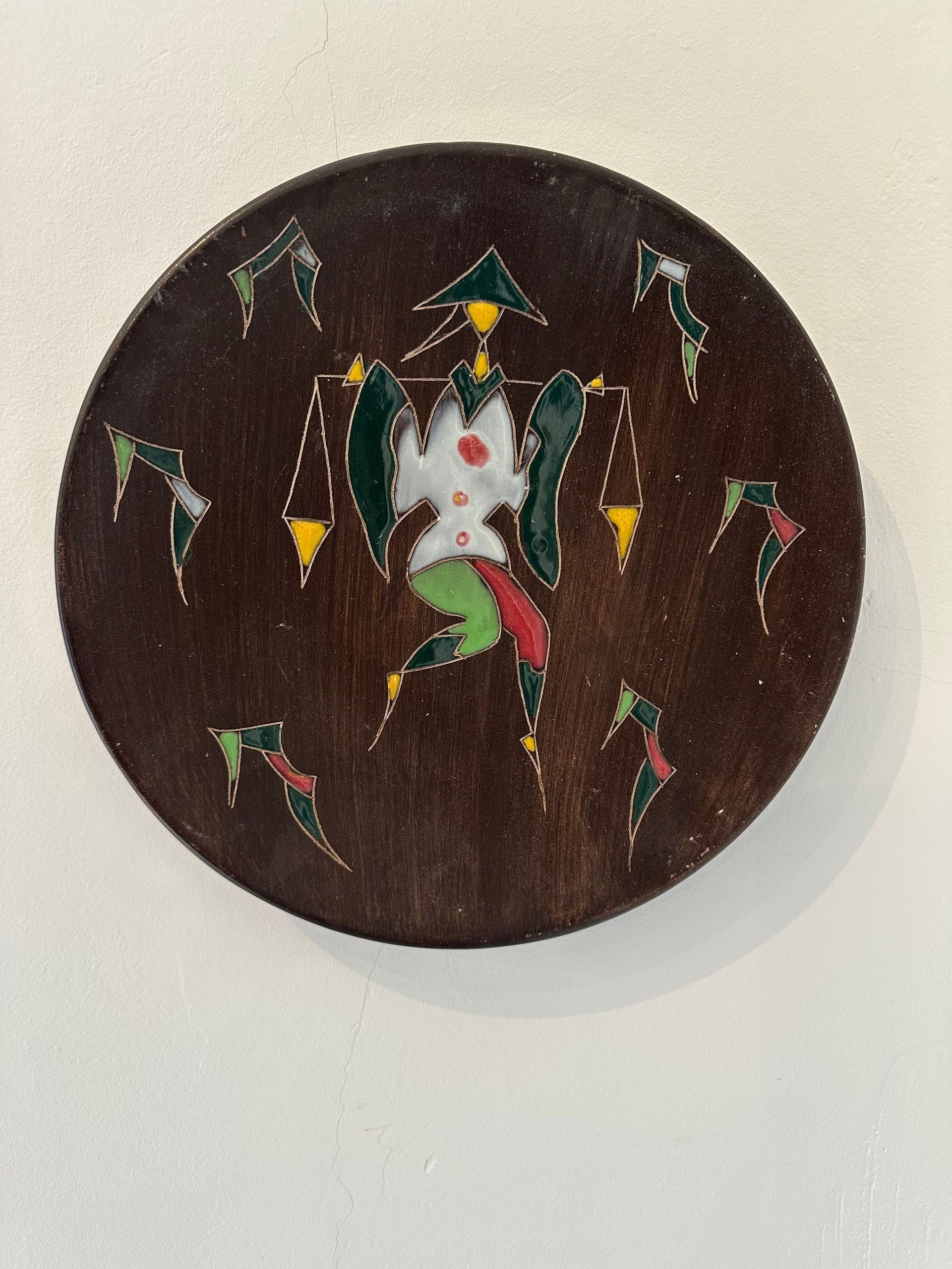 Plato redondo de madera oscura con una figura central en verde, blanco y rojo, rodeada de figuras similares.