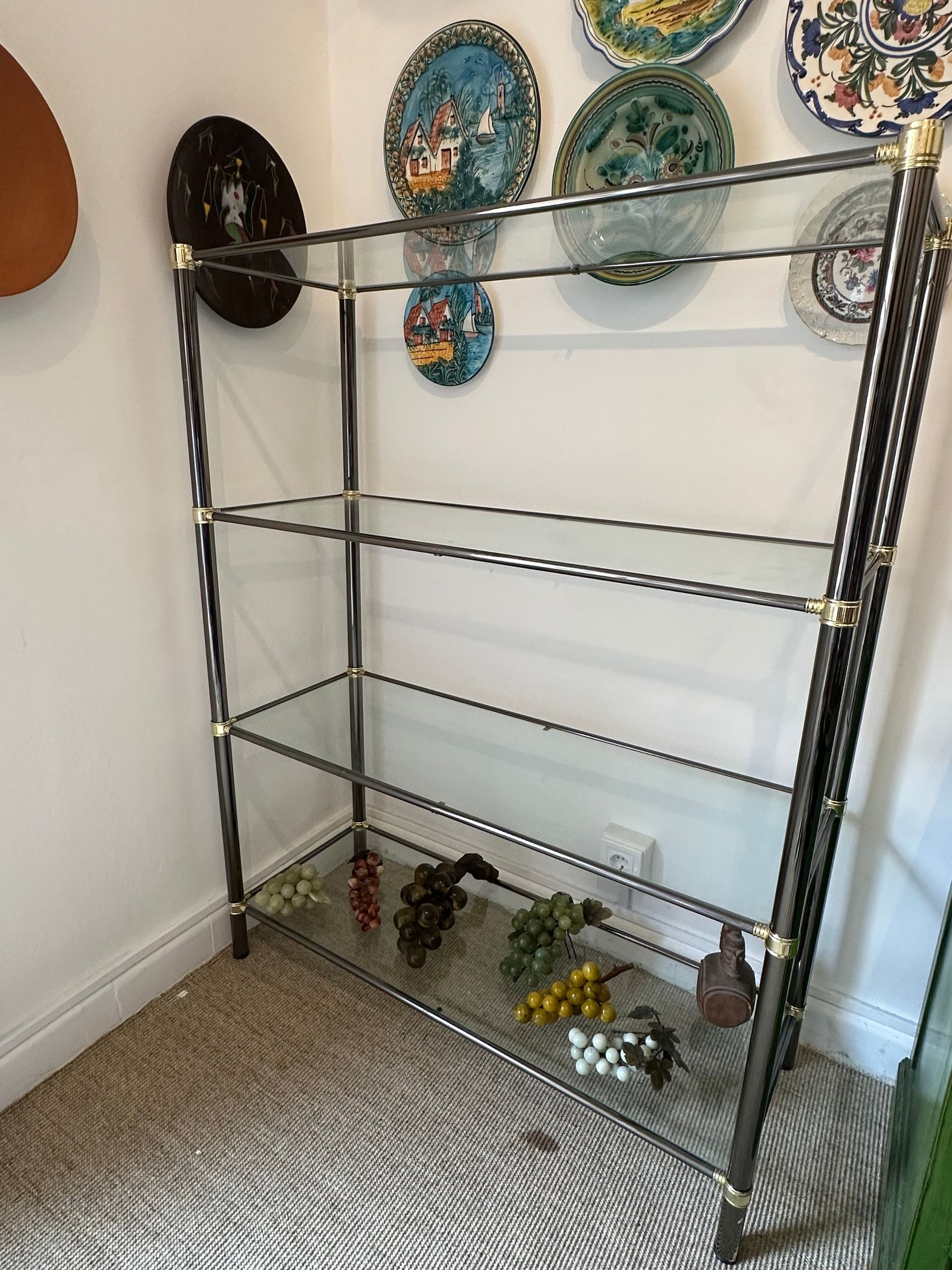Estantería de metal y vidrio con platos decorativos en la pared. Uvas artificiales en el estante inferior.