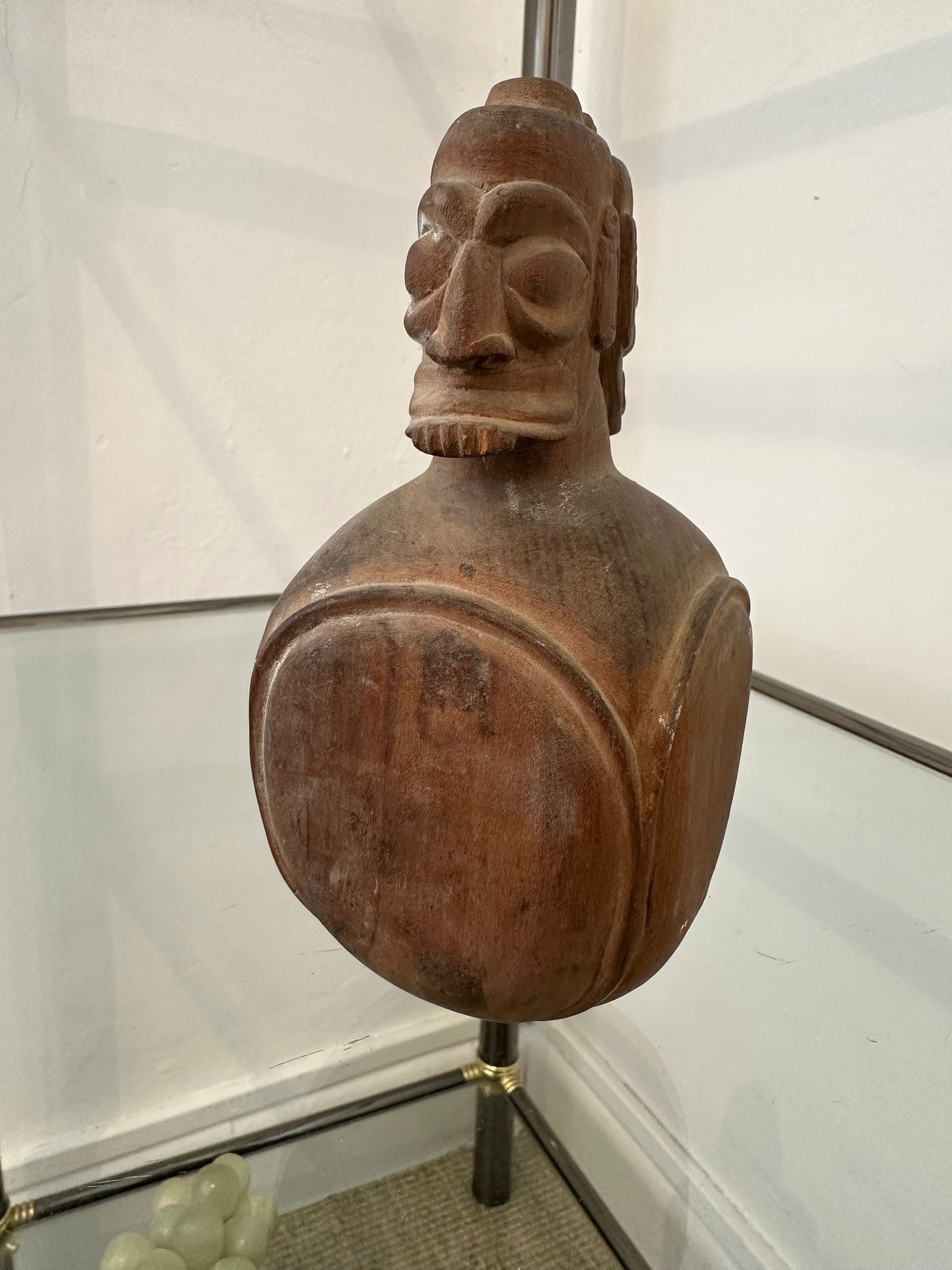 Escultura de madera de una cabeza sobre un cuerpo bulboso, posiblemente arte africano. Marrón, con rasgos faciales tallados.