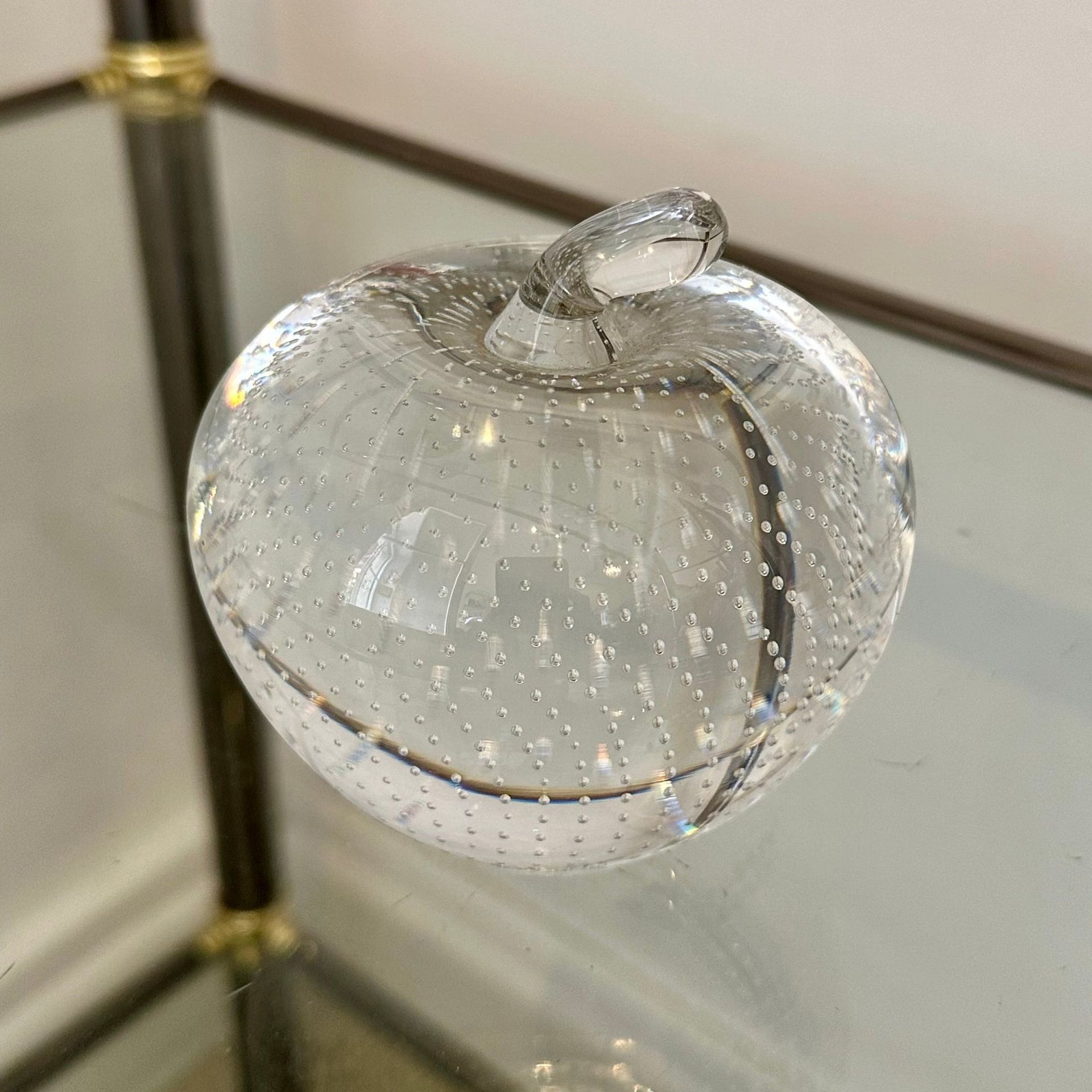 Manzana de vidrio transparente con tallo, apoyada sobre una superficie espejada.