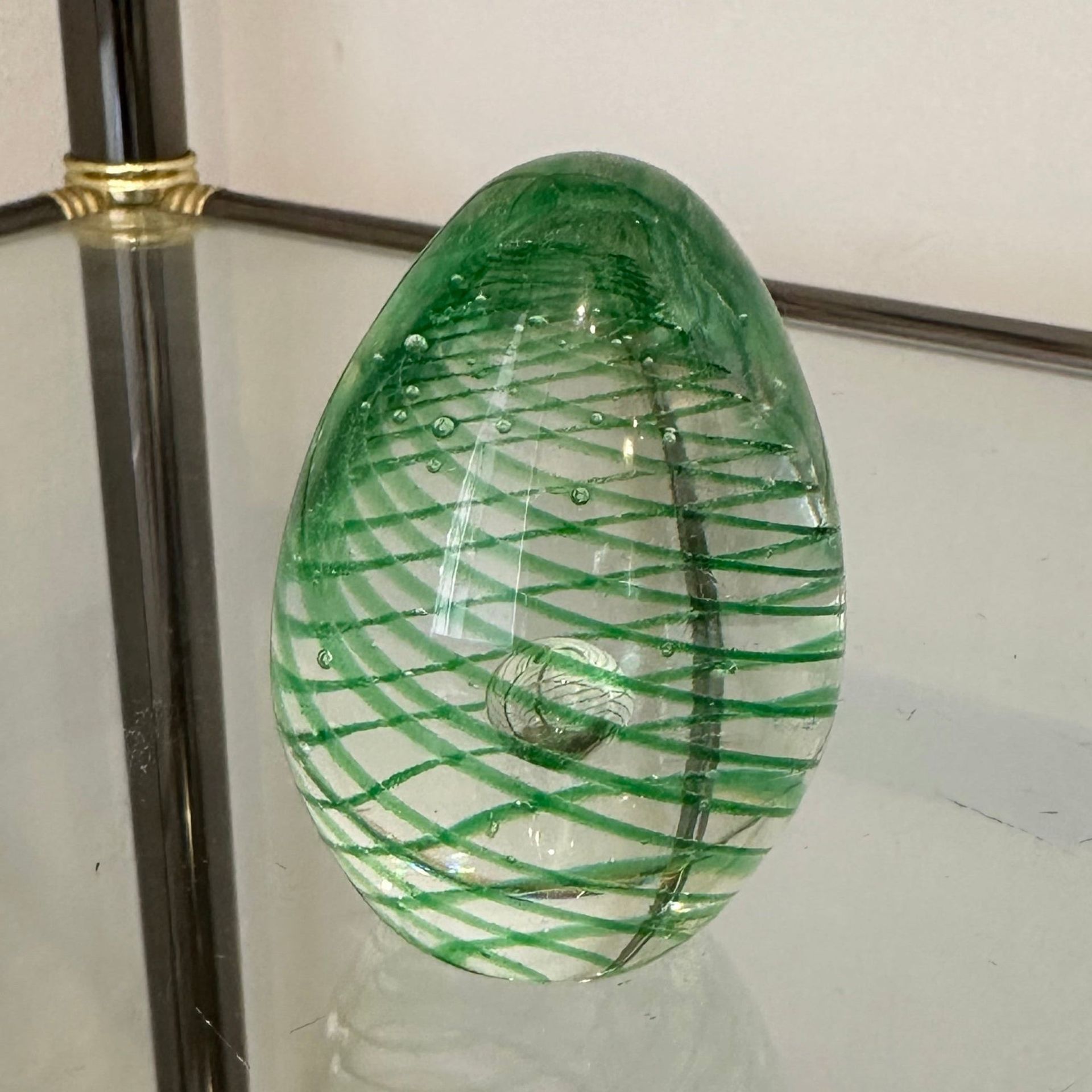 Huevo de vidrio verde con líneas verdes en espiral y burbujas de aire, apoyado sobre un estante de vidrio.