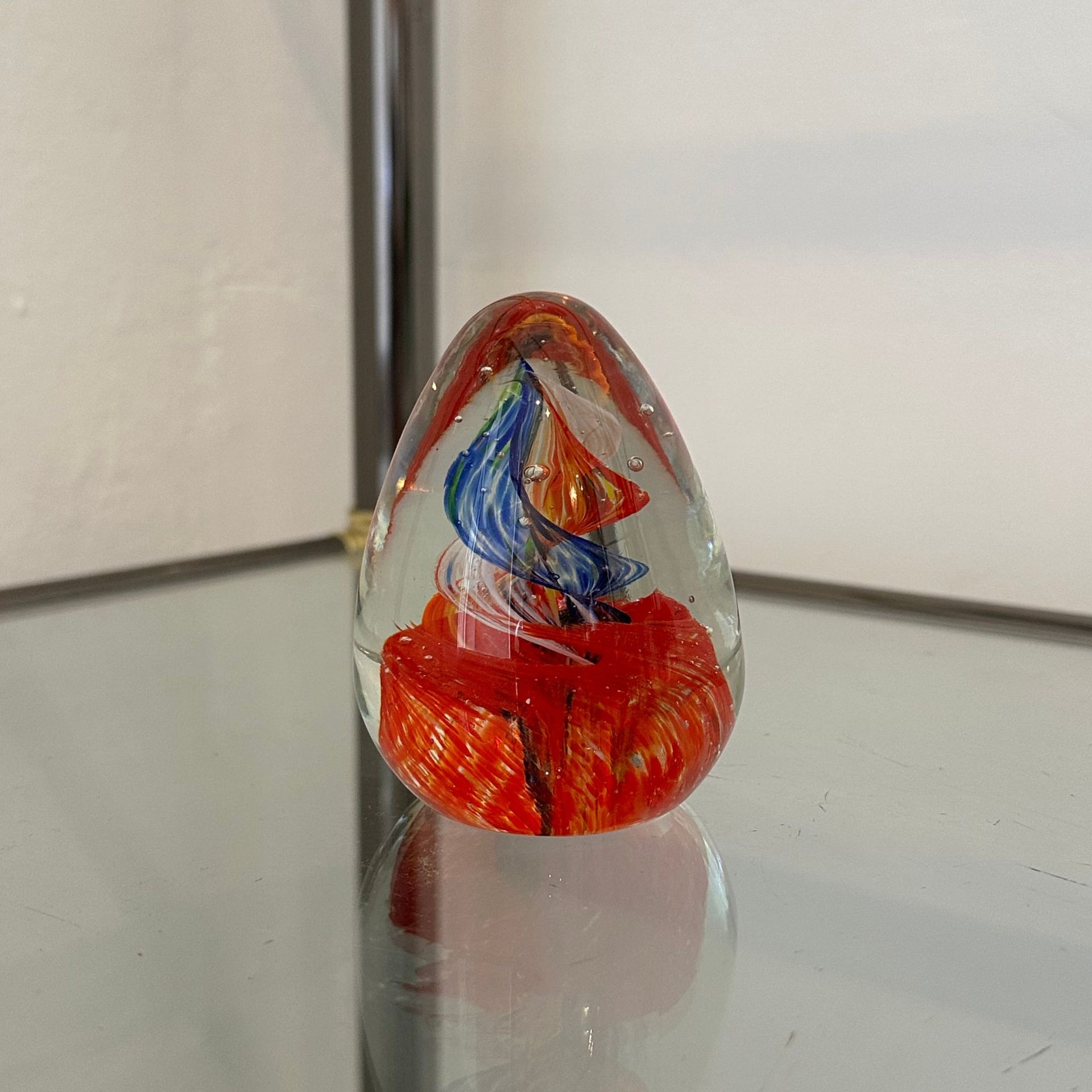 Pisapapeles en forma de huevo de cristal con espirales rojos y azules, sobre un estante con espejo.