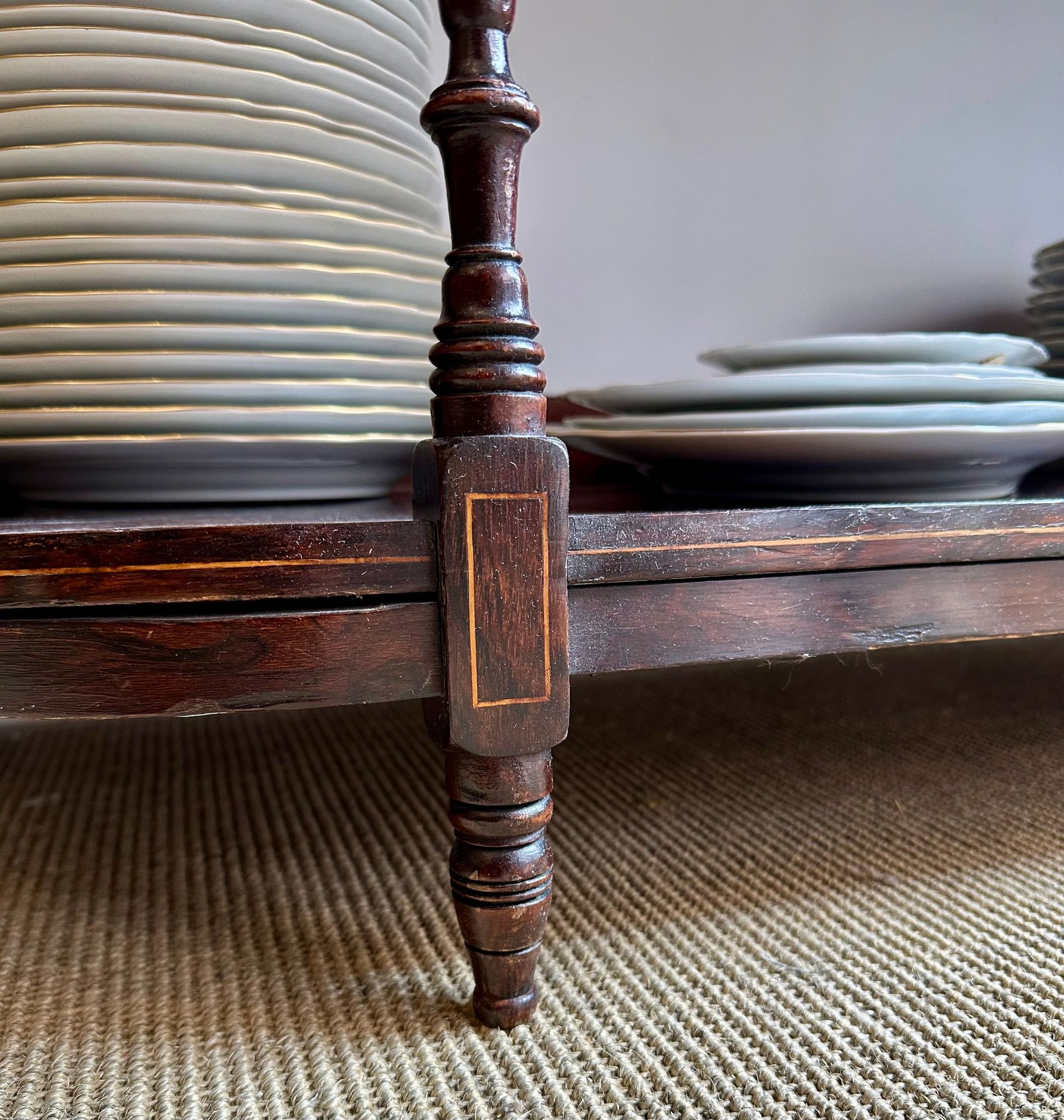 Primer plano de un estante de madera oscura con una pila de platos blancos. Se ve una pata de madera tallada.