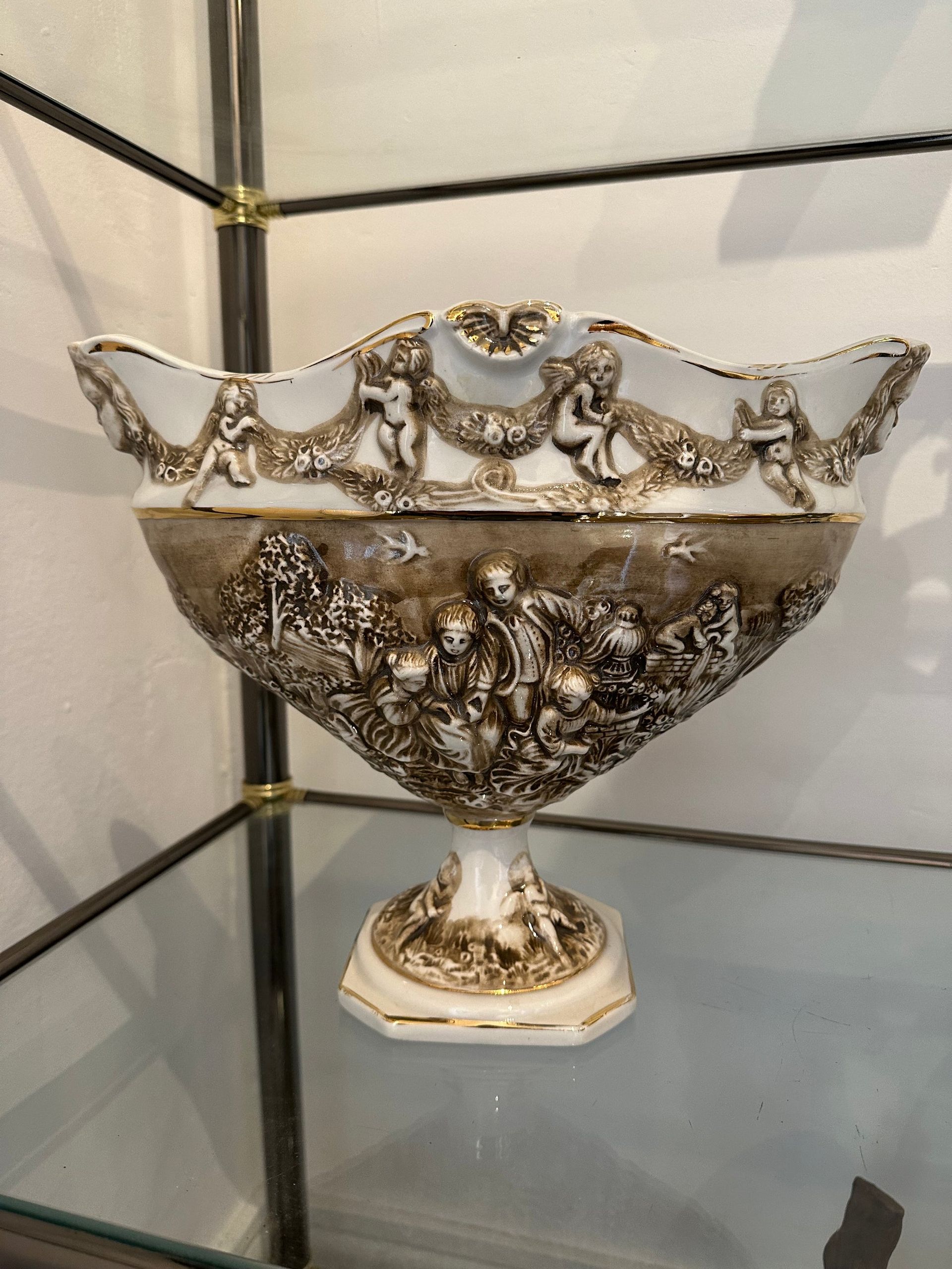 Cuenco de porcelana blanca y dorada adornado con figuras y querubines en un estante.