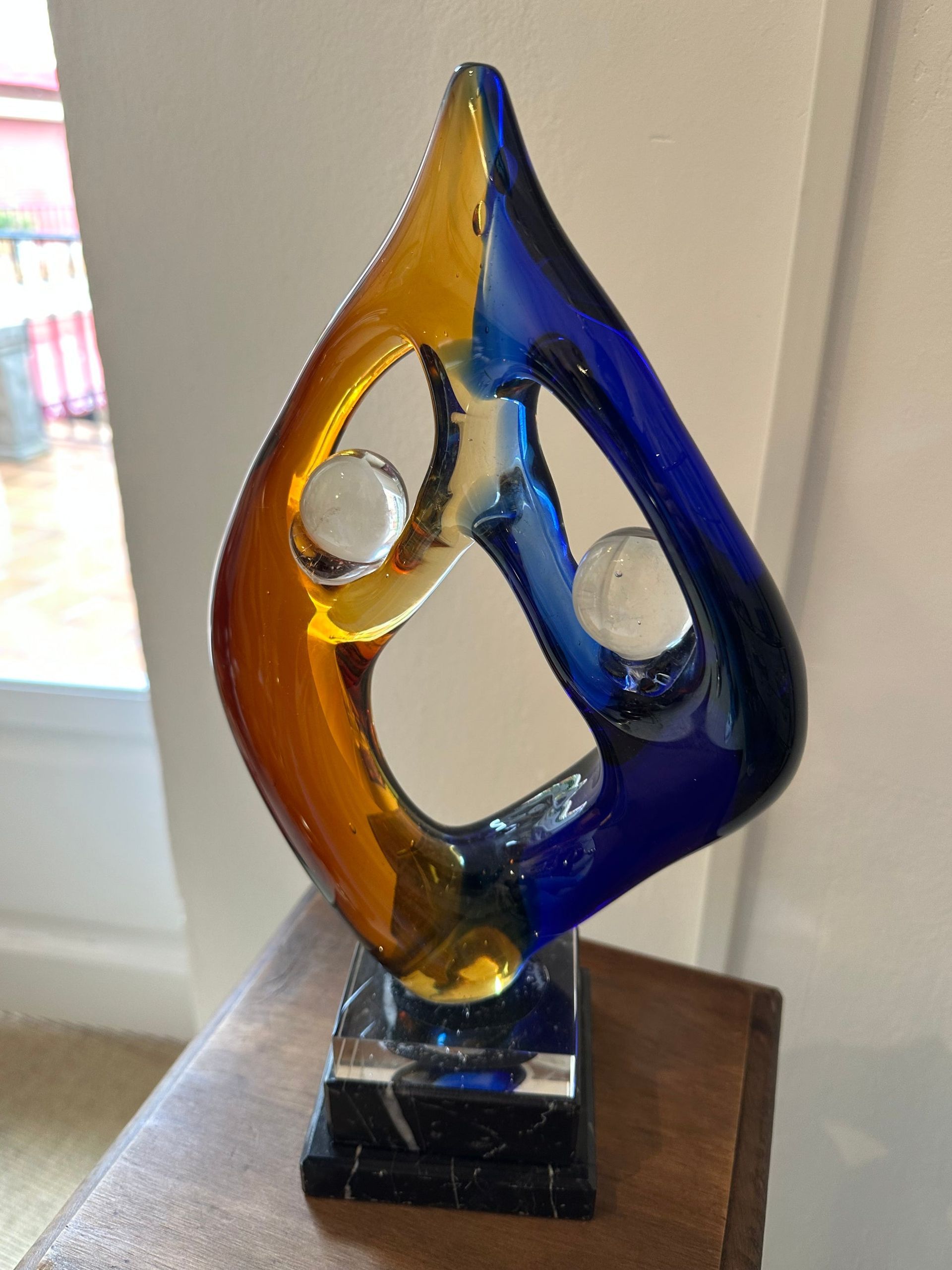 Escultura de vidrio, forma de lágrima azul y amarilla, dos esferas blancas, base negra.