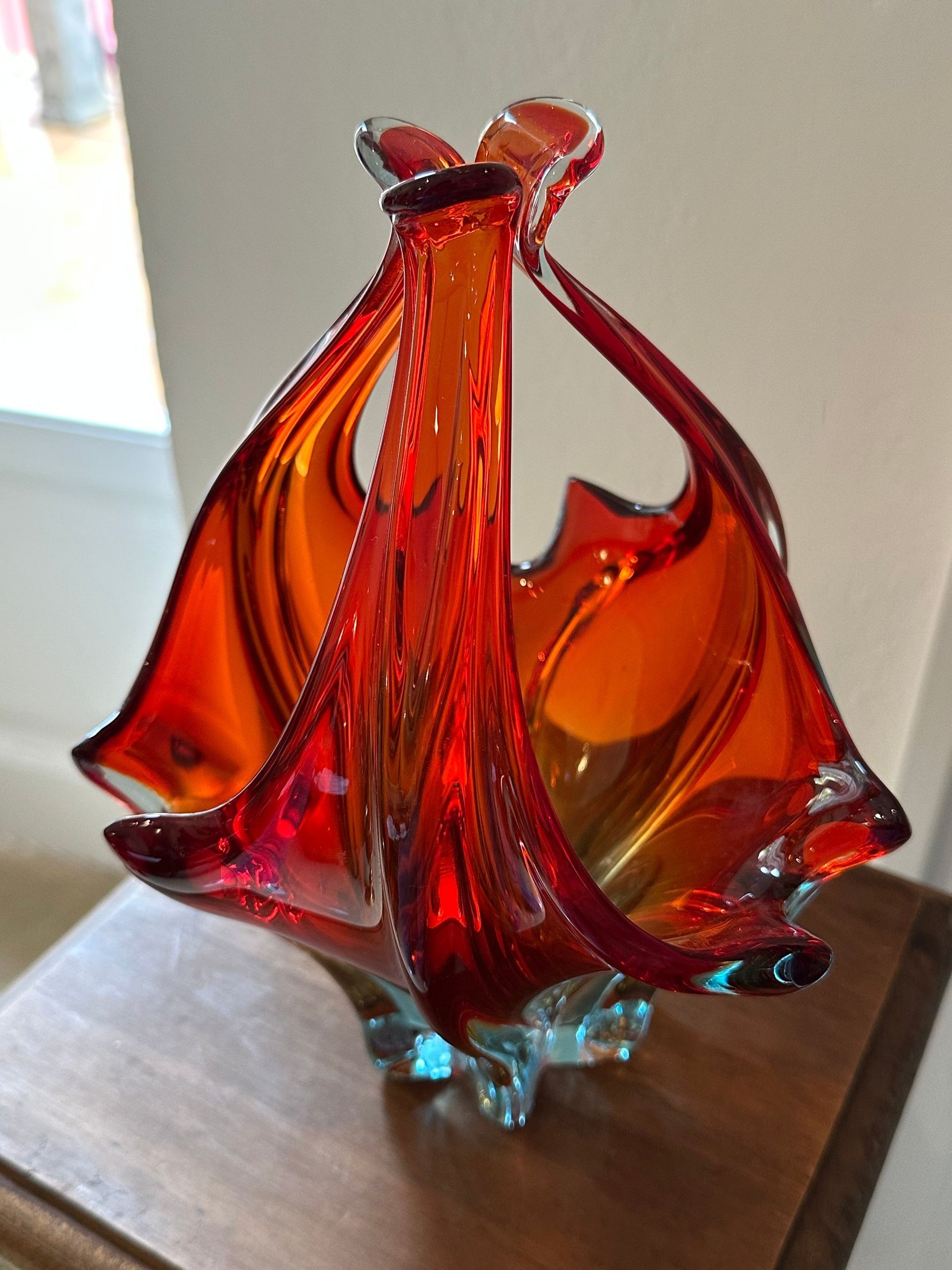 Cesta de cristal de color naranja y rojo con base de color azul transparente, sobre superficie de madera.