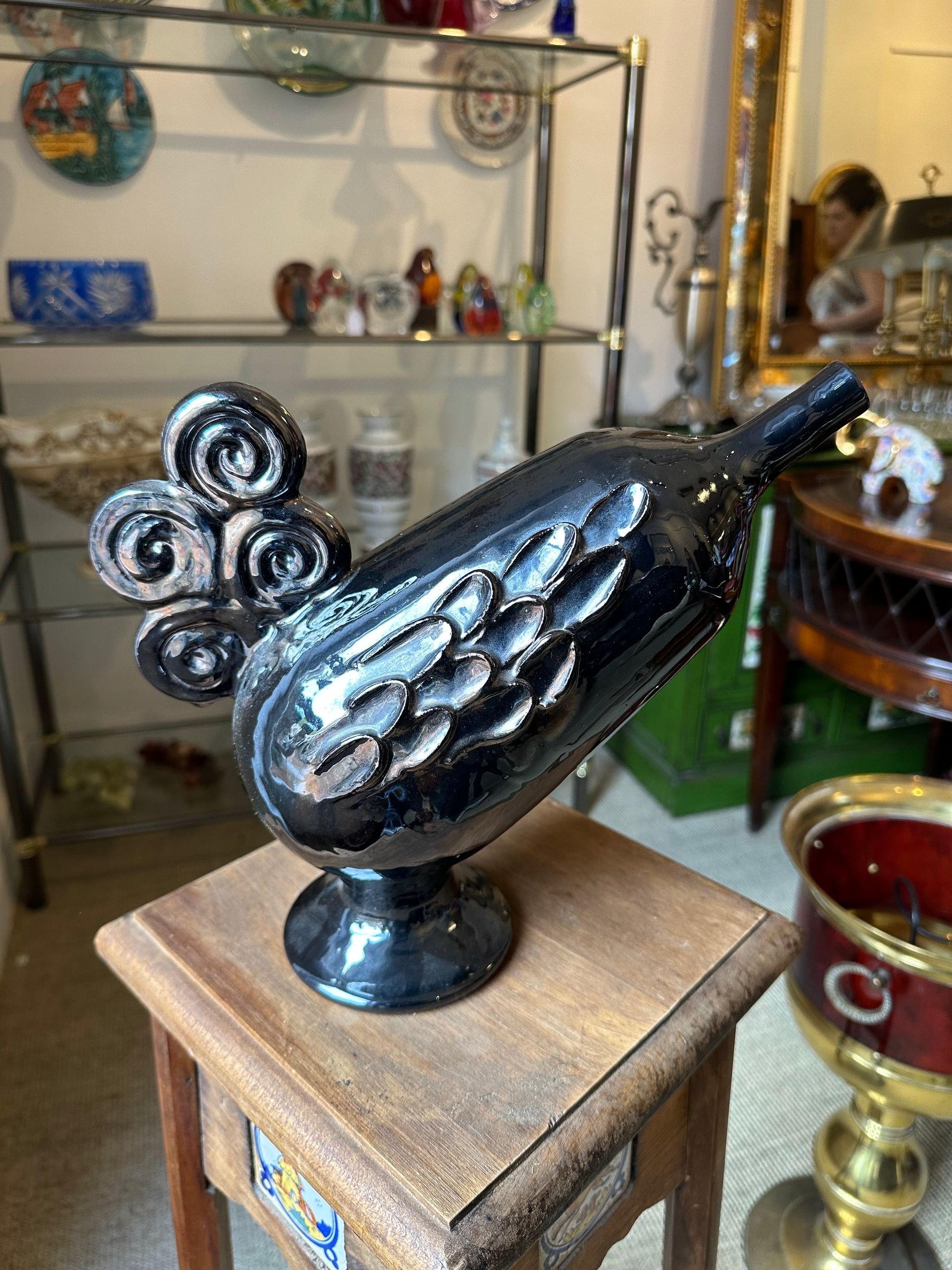 Estatua de gallo de cerámica negra sobre una pequeña mesa de madera, expuesta en una tienda junto a otros objetos.