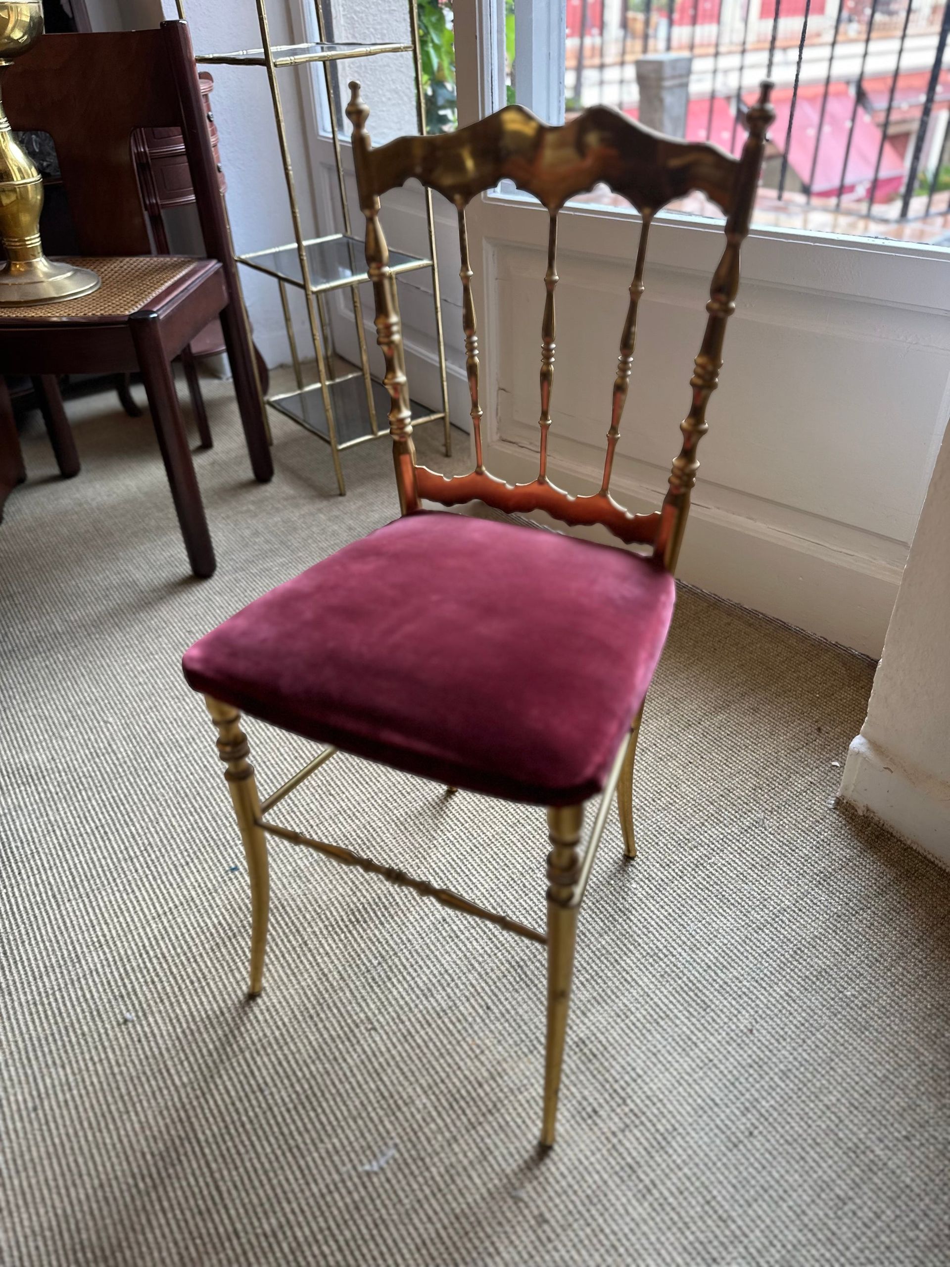 Silla con marco dorado y asiento de terciopelo burdeos frente a una ventana; también hay otros muebles y una alfombra.