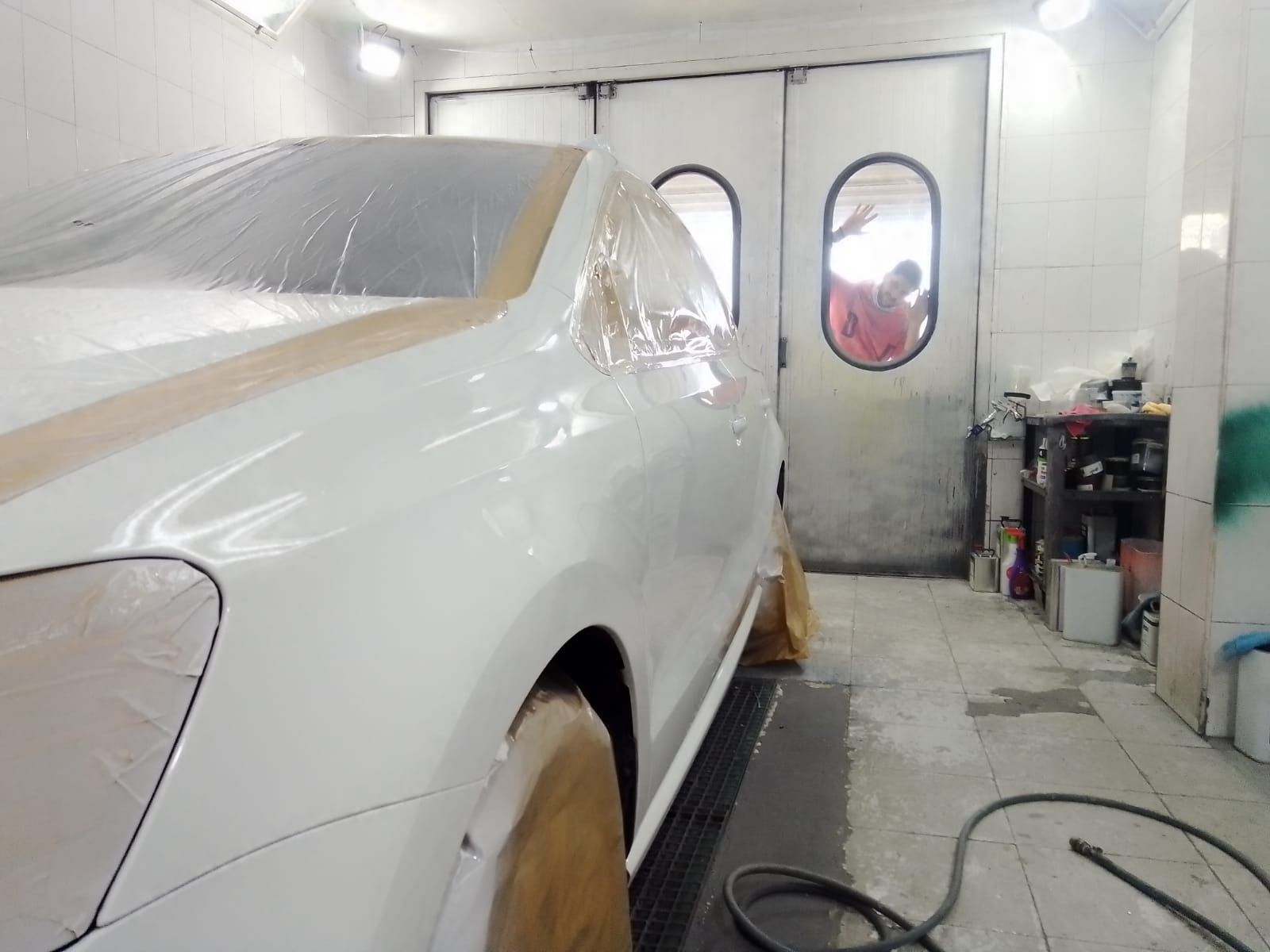 Coche blanco en una cabina de pintura, parcialmente tapado para pintar. Vista interior con una persona visible a través de una puerta.