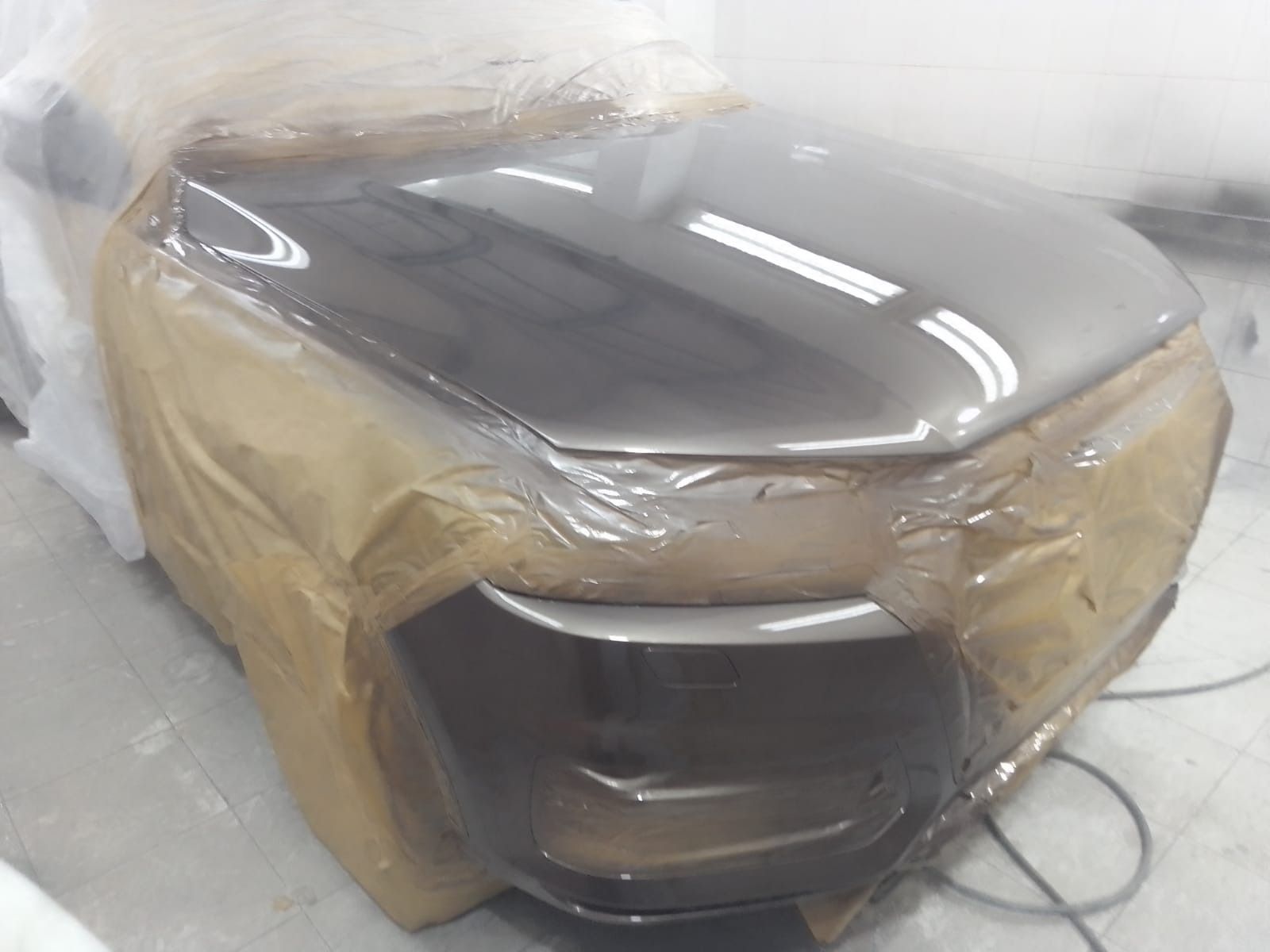 Capó del coche en proceso de pintura; color gris oscuro con cinta de enmascarar y envoltura plástica protegiendo otras partes.