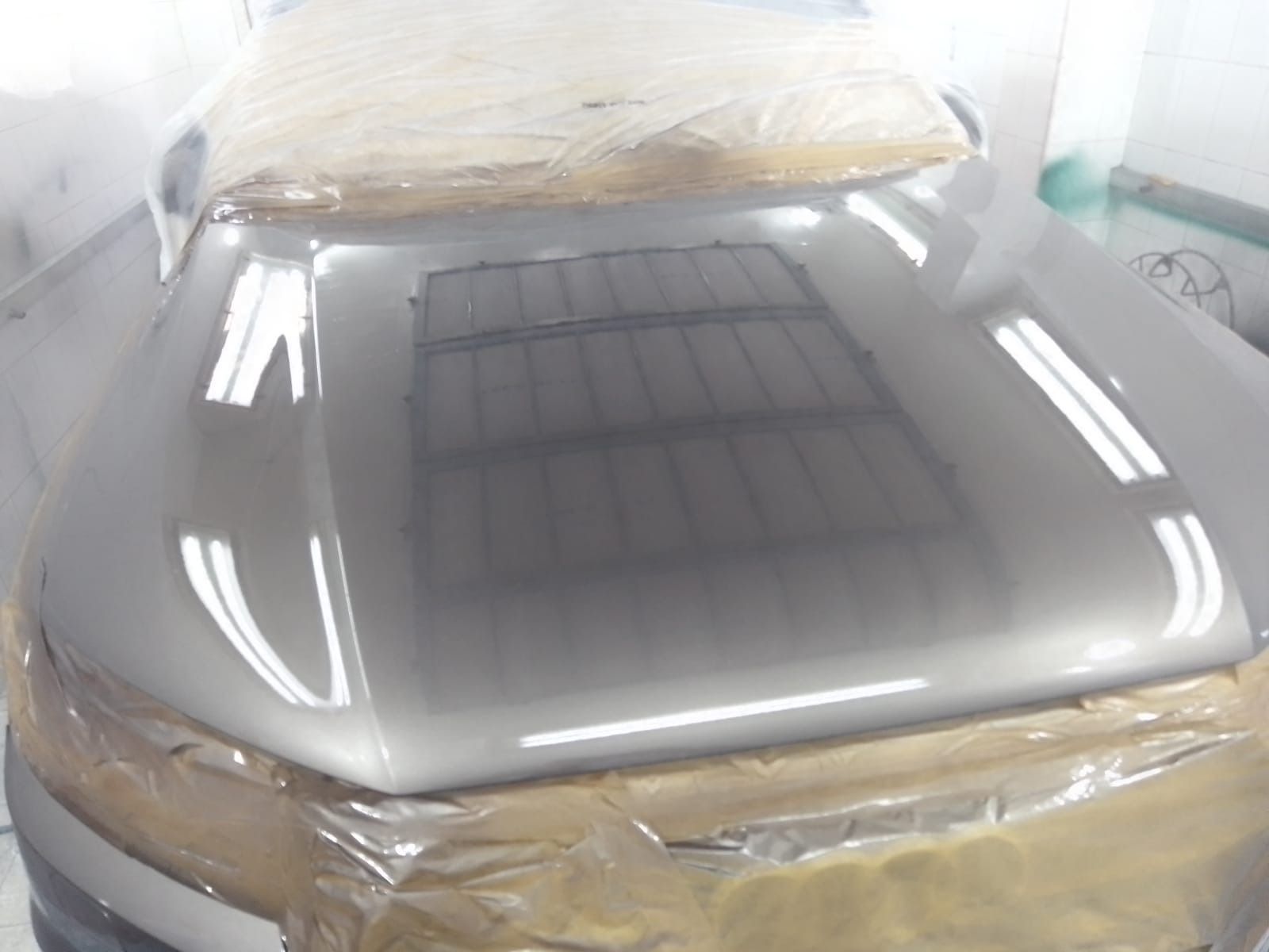 El capó de un coche, recién pintado de color plata, refleja las luces del techo de un taller, cubierto de plástico para su protección.