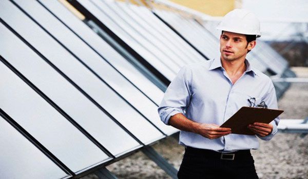 Un hombre que lleva un casco sostiene un portapapeles delante de un panel solar.