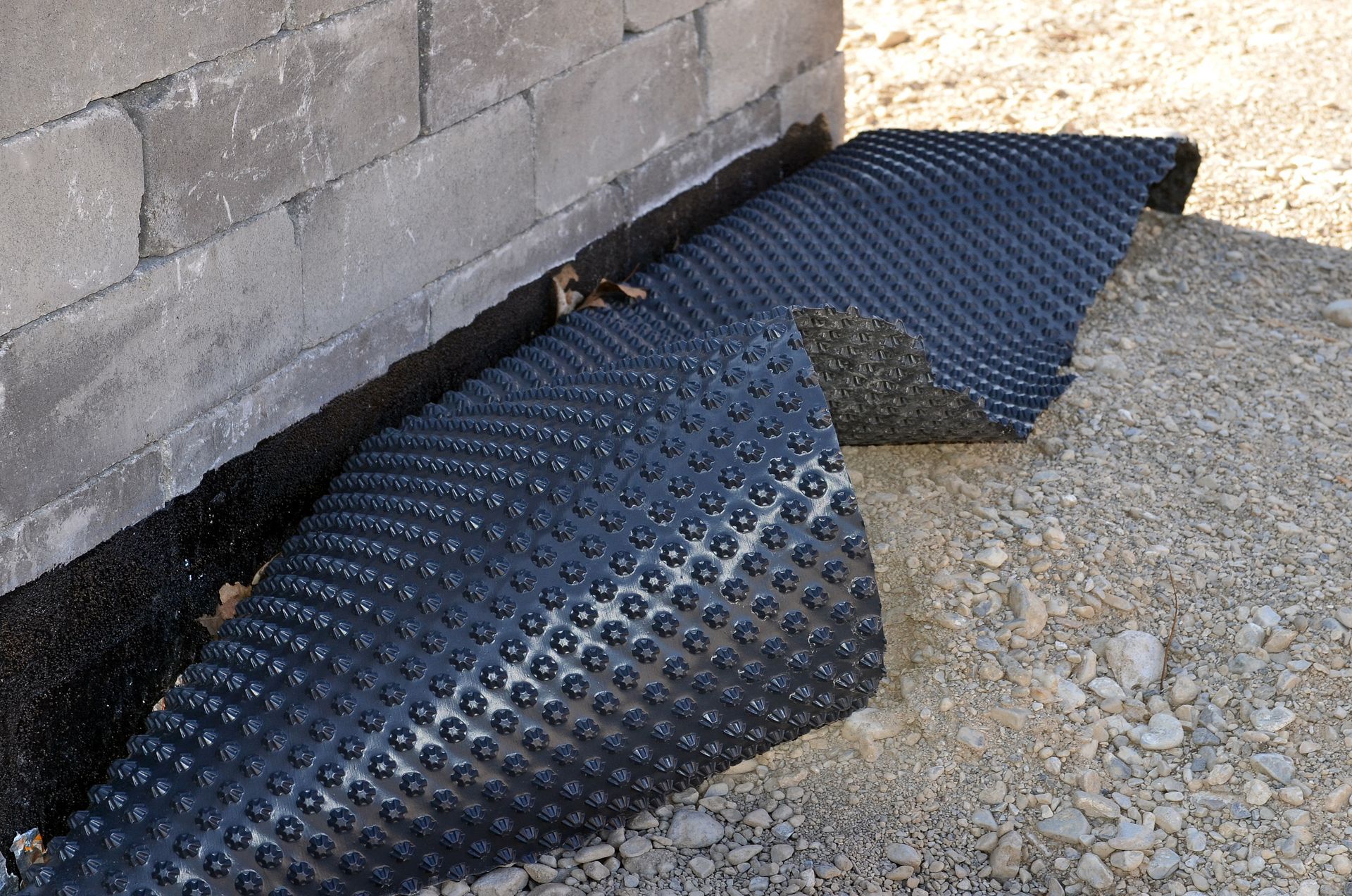 Eine Rolle schwarzer Drainagematten liegt auf dem Boden neben einer Backsteinmauer.