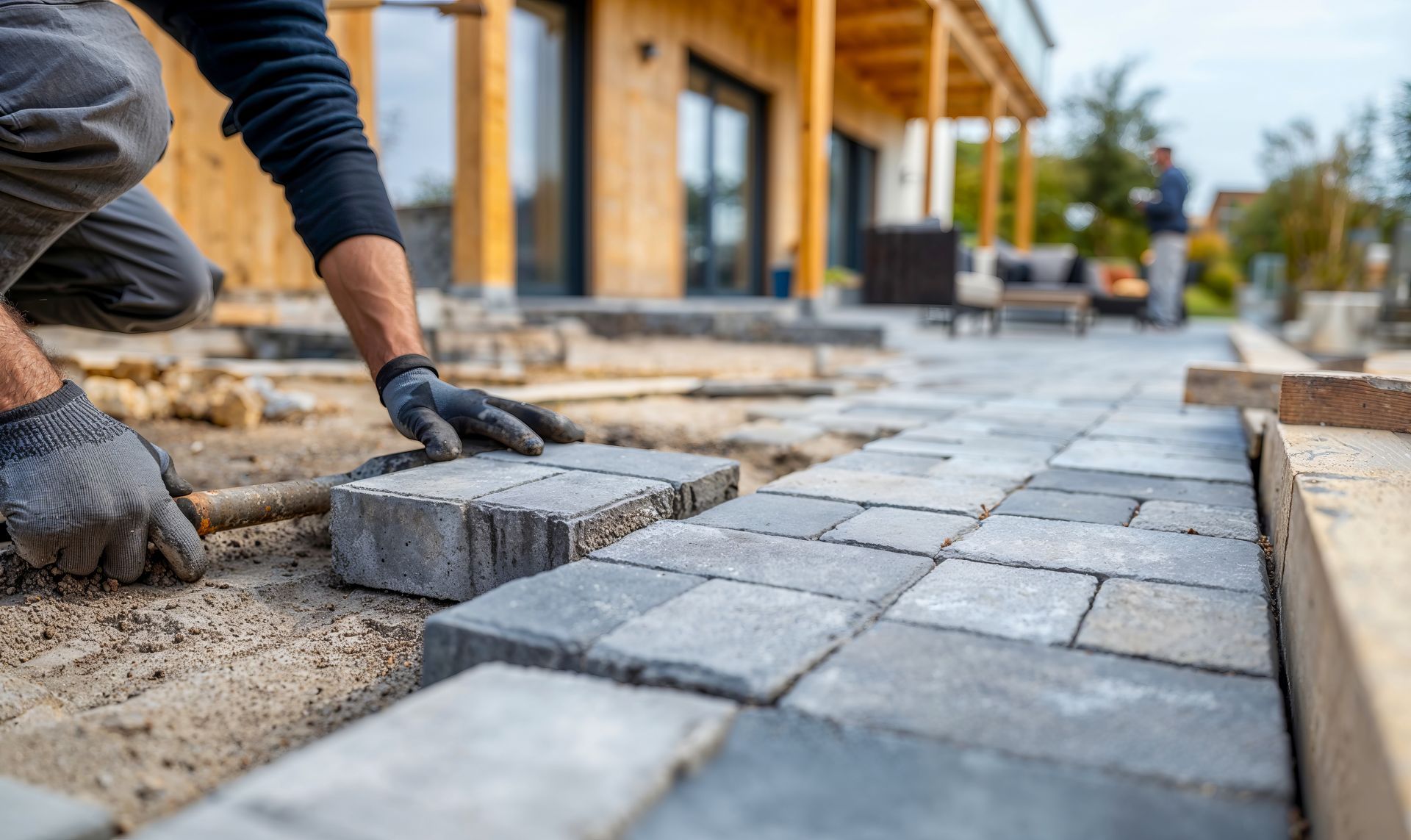 Un ouvrier pose des pavés gris le long d'un trottoir, à côté d'un bâtiment en construction.