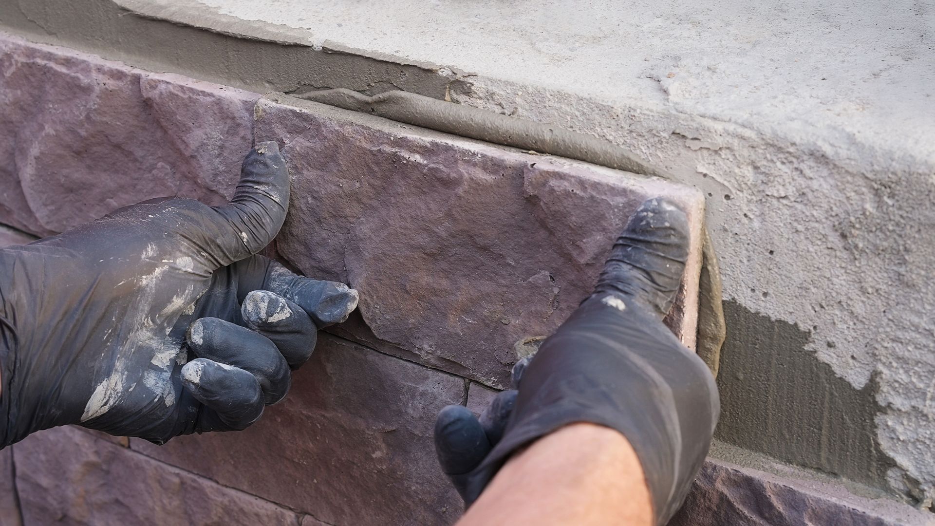 Des mains gantées ajustent un panneau de placage en pierre sur un mur extérieur.