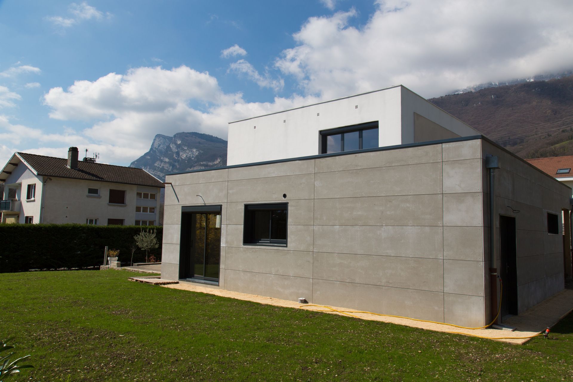 Maison moderne en béton sur une pelouse avec des montagnes en arrière-plan.