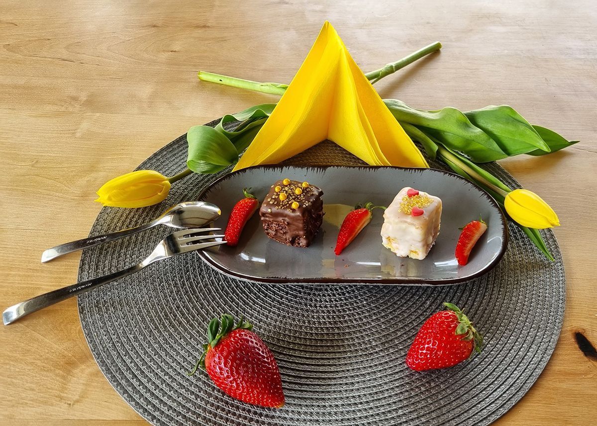 Ein Gourmet-Dessert auf schwarzem Teller mit gelber Garnitur und Erdbeeren auf einem gewebten Platzdeckchen.