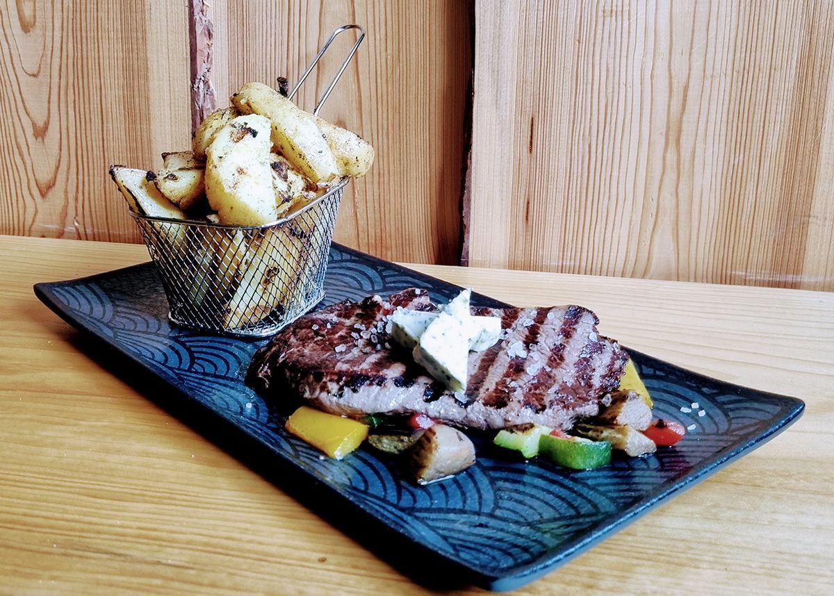 Gegrilltes Steak mit Gemüse auf einer schwarzen Platte, daneben Brot in einem Metallbecher auf einem Holztisch.