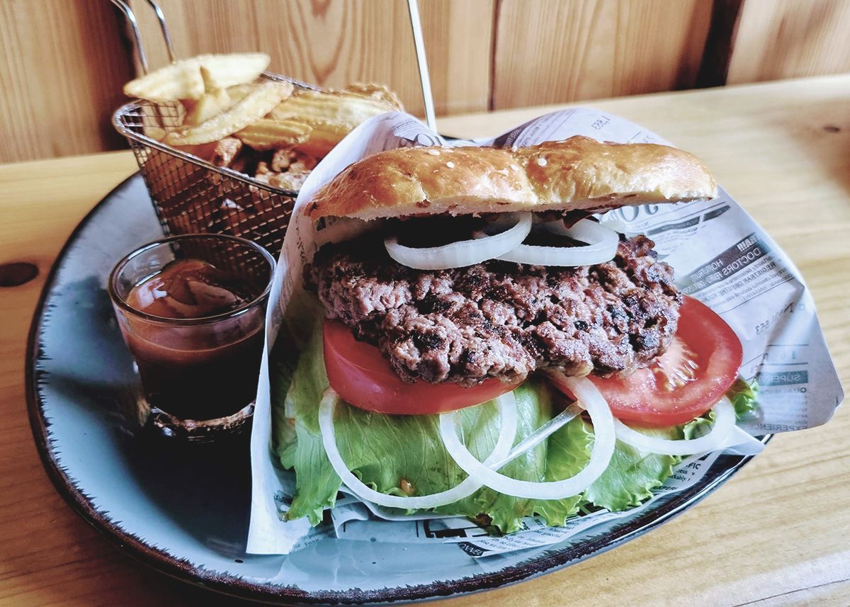 Burger mit Tomaten und Salat auf einem Teller, serviert mit Pommes frites und einem Schälchen Soße auf einem Holztisch.