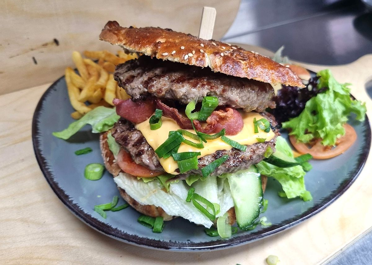 Ein hoher, gestapelter Burger mit Salat, Käse und Pommes frites auf einem dunklen Teller.