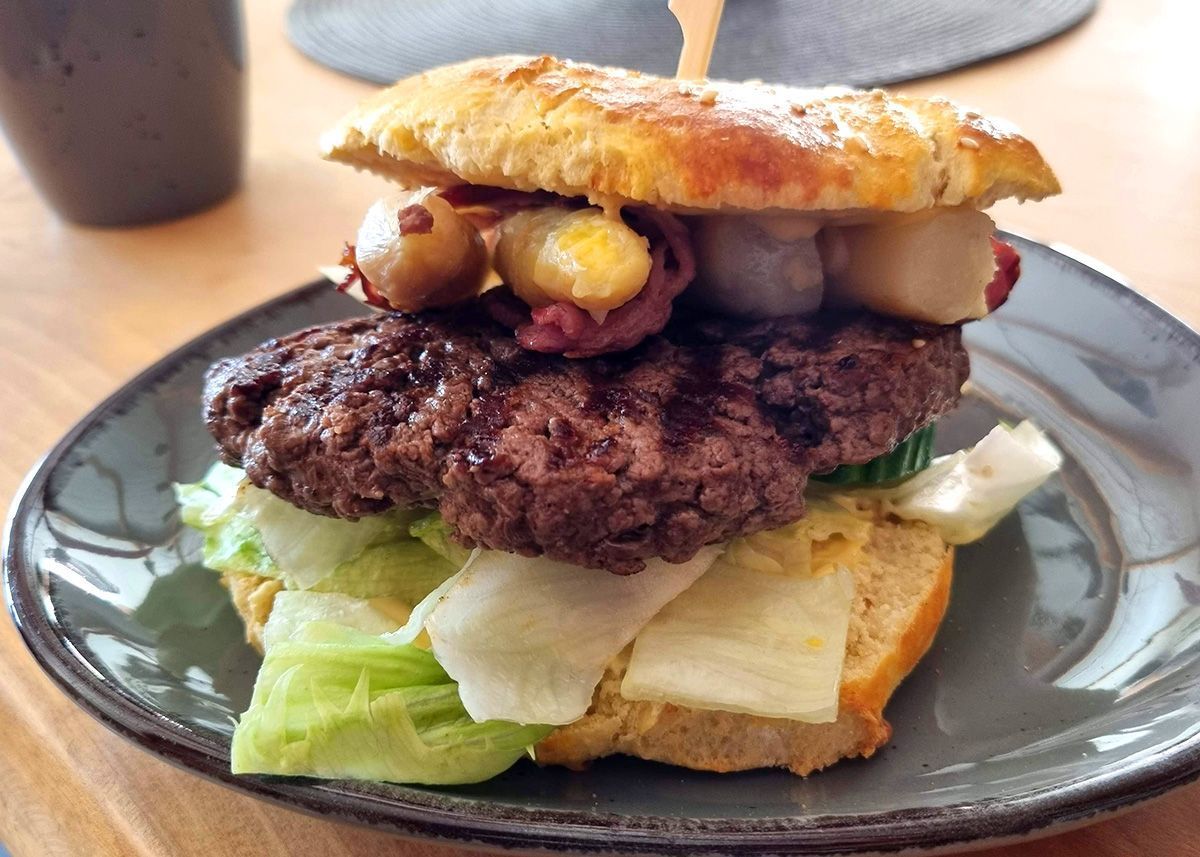Nahaufnahme eines Cheeseburgers auf einem dunklen Teller mit Salat, Zwiebeln, Speck und einem gerösteten Brötchen.
