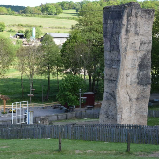 Hohe, graue Kletterwand in einem grasbewachsenen Park mit Bäumen und Hügeln im Hintergrund.