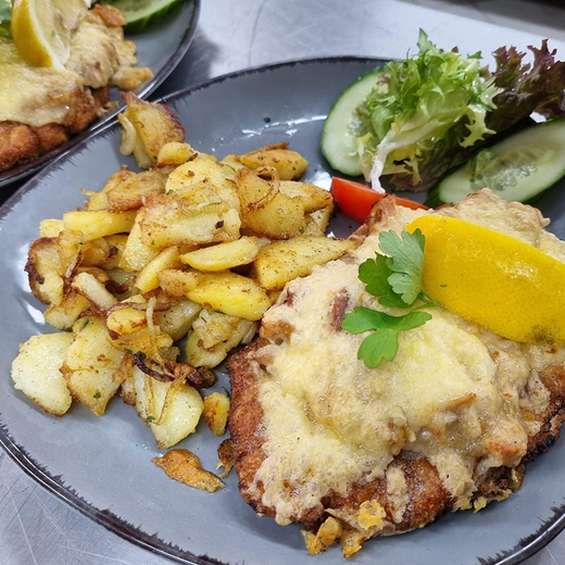 Teller mit paniertem Fisch, Bratkartoffeln, Gurkensalat und Zitronenspalte auf einem dunklen Teller