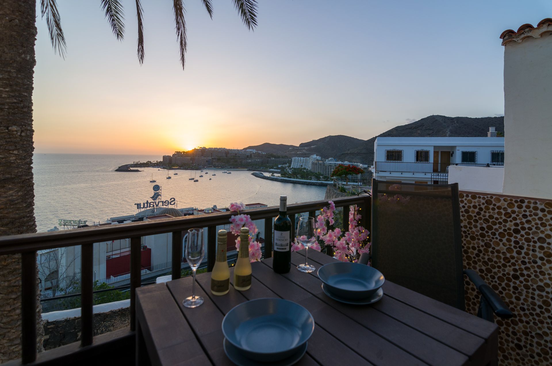 Una mesa con platos y una botella de vino sobre ella con vistas al océano al atardecer.