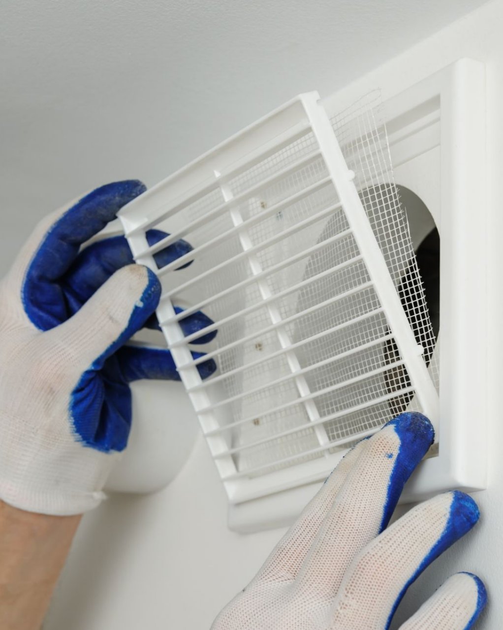 Des mains gantées installent une grille d'aération blanche sur un mur.