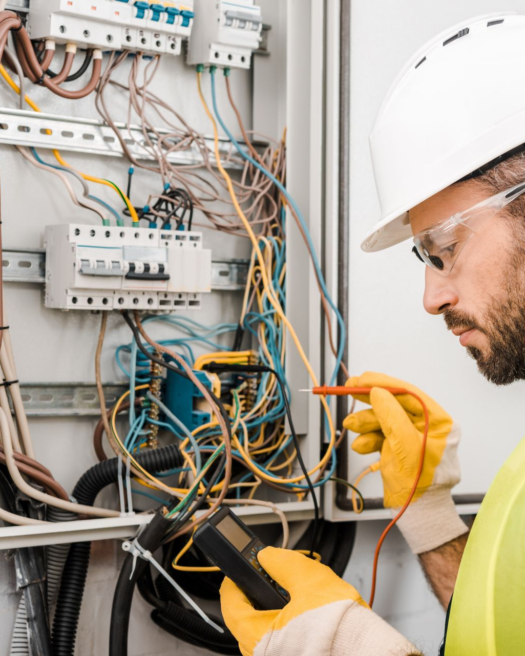 Un électricien portant un casque et des lunettes de sécurité utilise un multimètre sur le câblage électrique d'un tableau électrique.