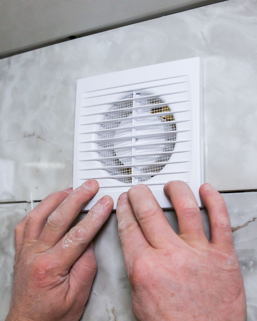 Des mains installent un ventilateur blanc sur un mur en marbre.