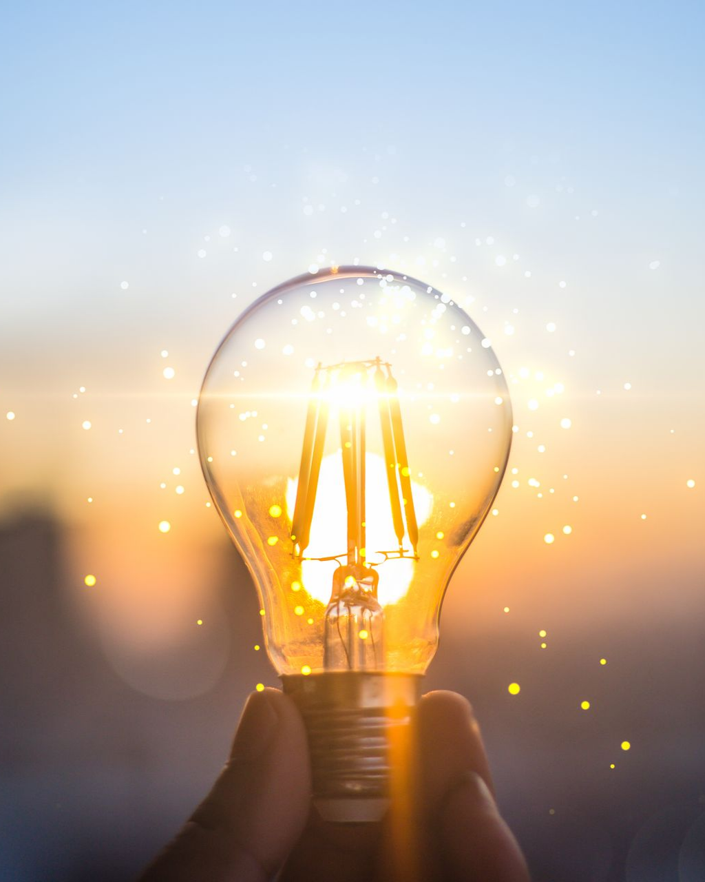 Une main tenant une ampoule allumée sur fond de coucher de soleil doré et flou.