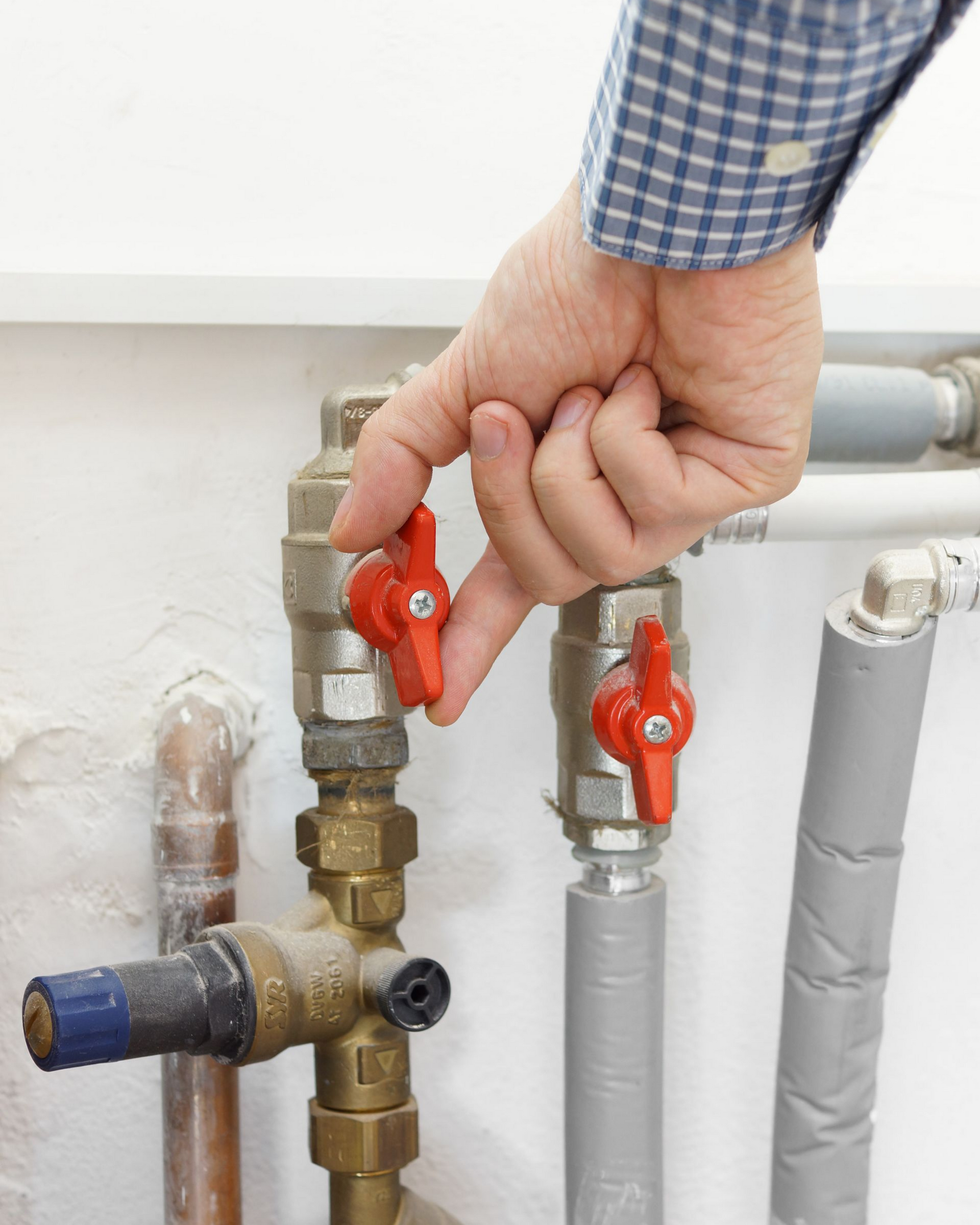 Une main actionne une vanne rouge sur des tuyaux de plomberie, à l'intérieur.